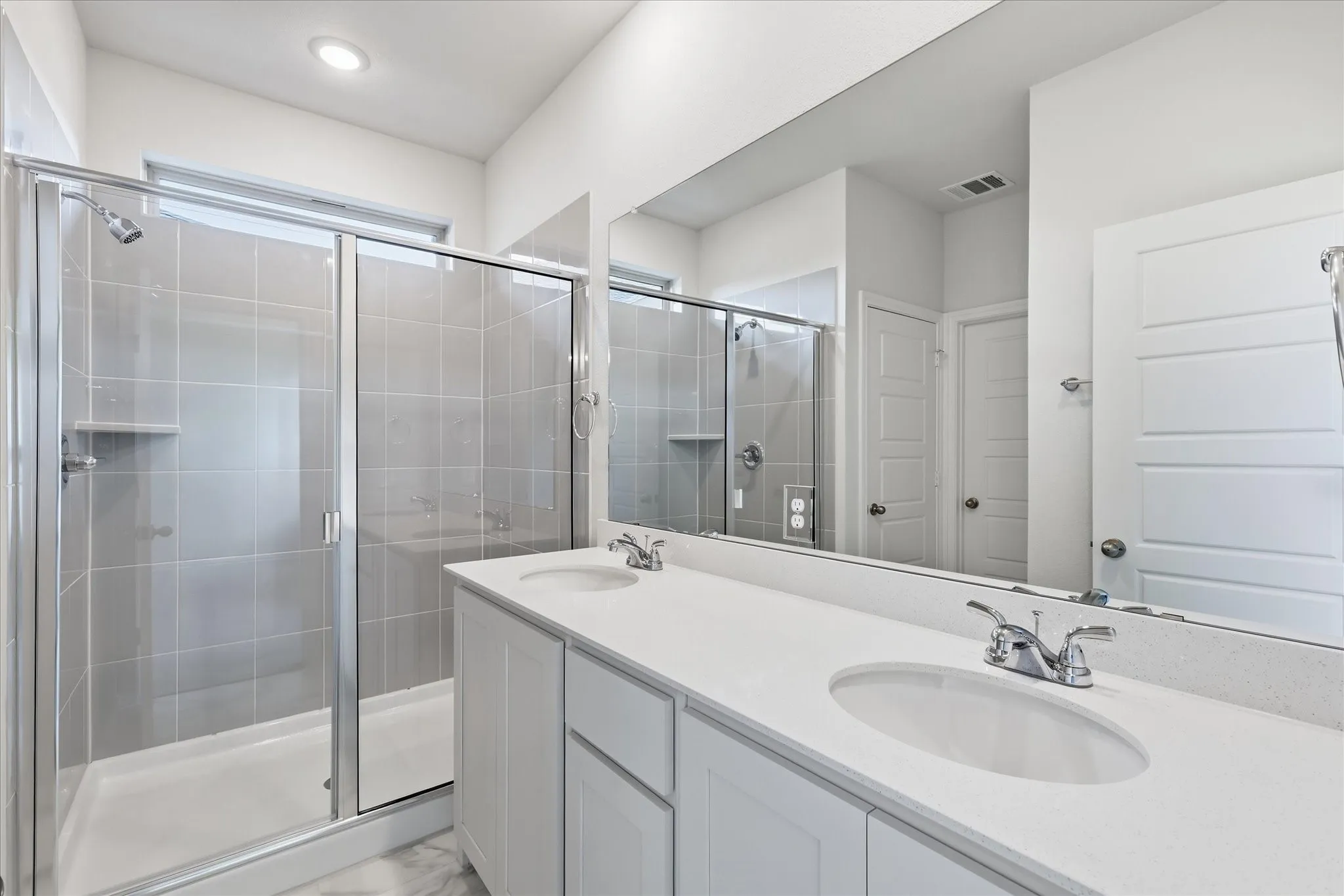 Bathroom featuring a dual vanity with white cabinetry and integrated sinks, a wide mirror, and a glass-enclosed shower with light gray tiled walls