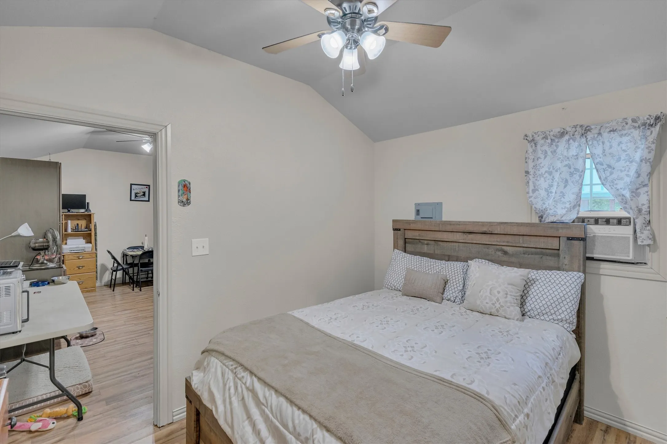 Vaulted ceiling room featuring a ceiling fan with light fixture, wood-finish flooring, and a window with an installed air conditioning unit