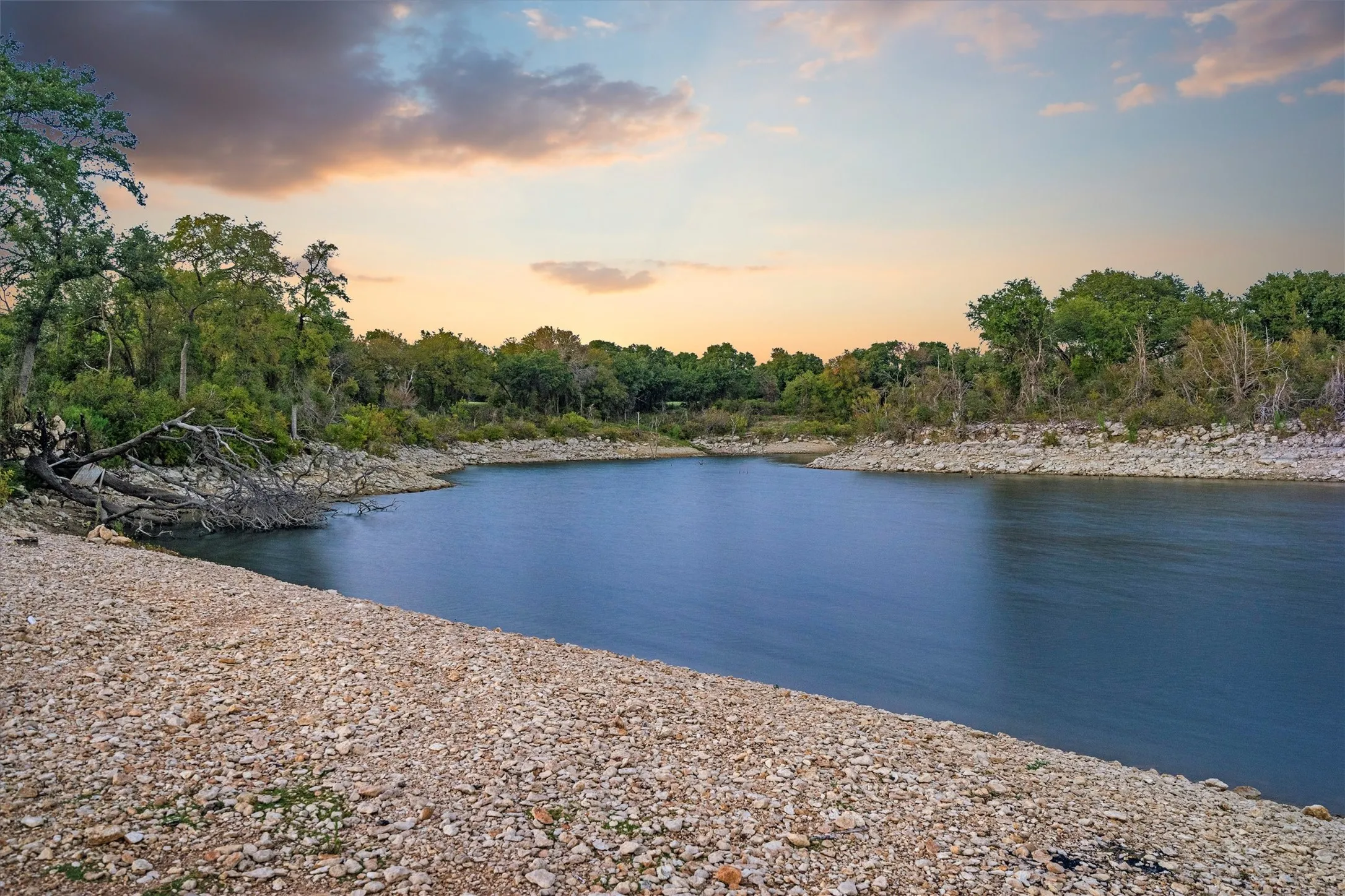 Expansive waterfront shoreline featuring a rocky beach, calm water, and mature trees lining the distant bank