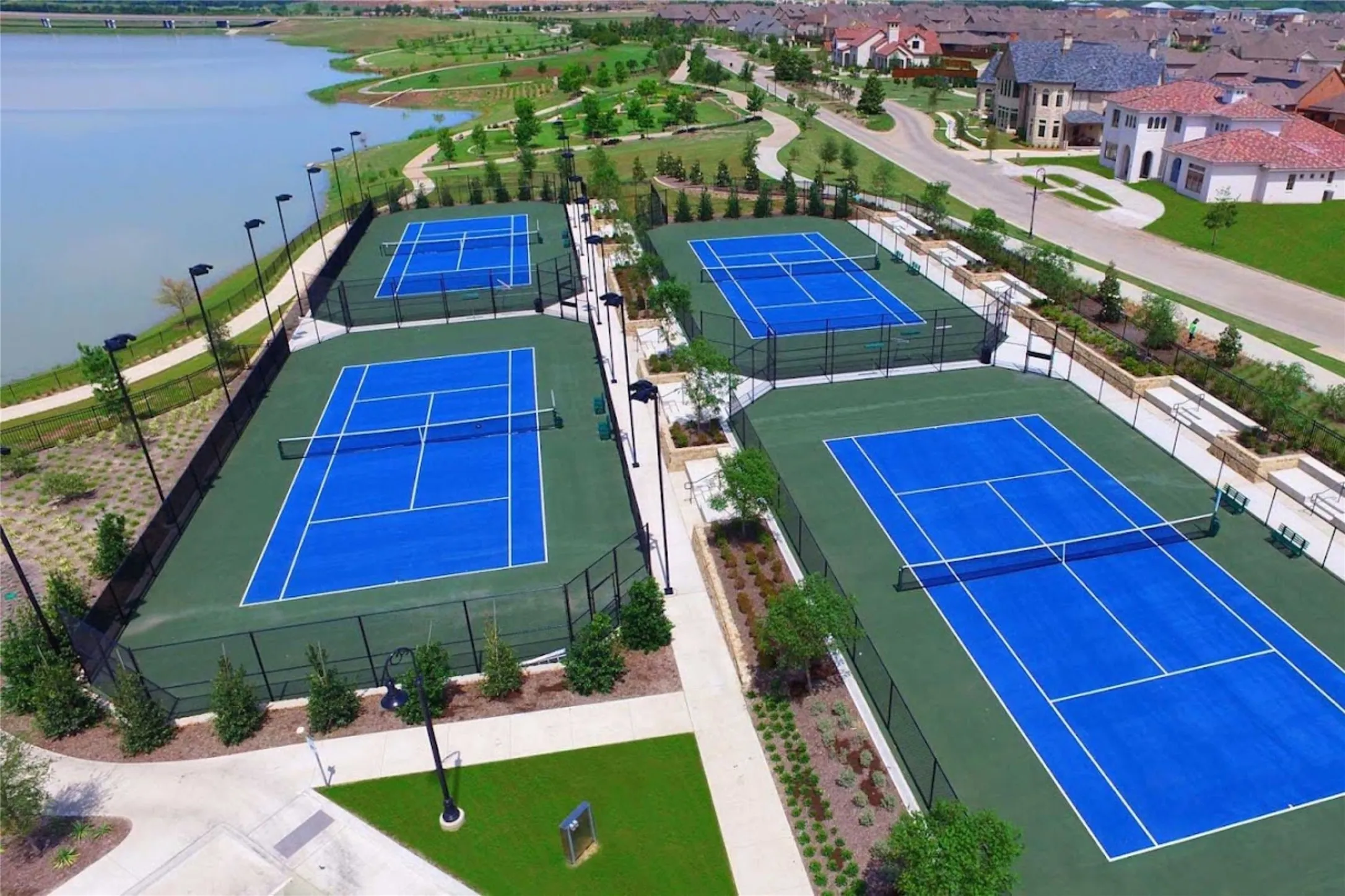 Multiple tennis courts with blue playing surfaces and green surrounds, illuminated by numerous light poles