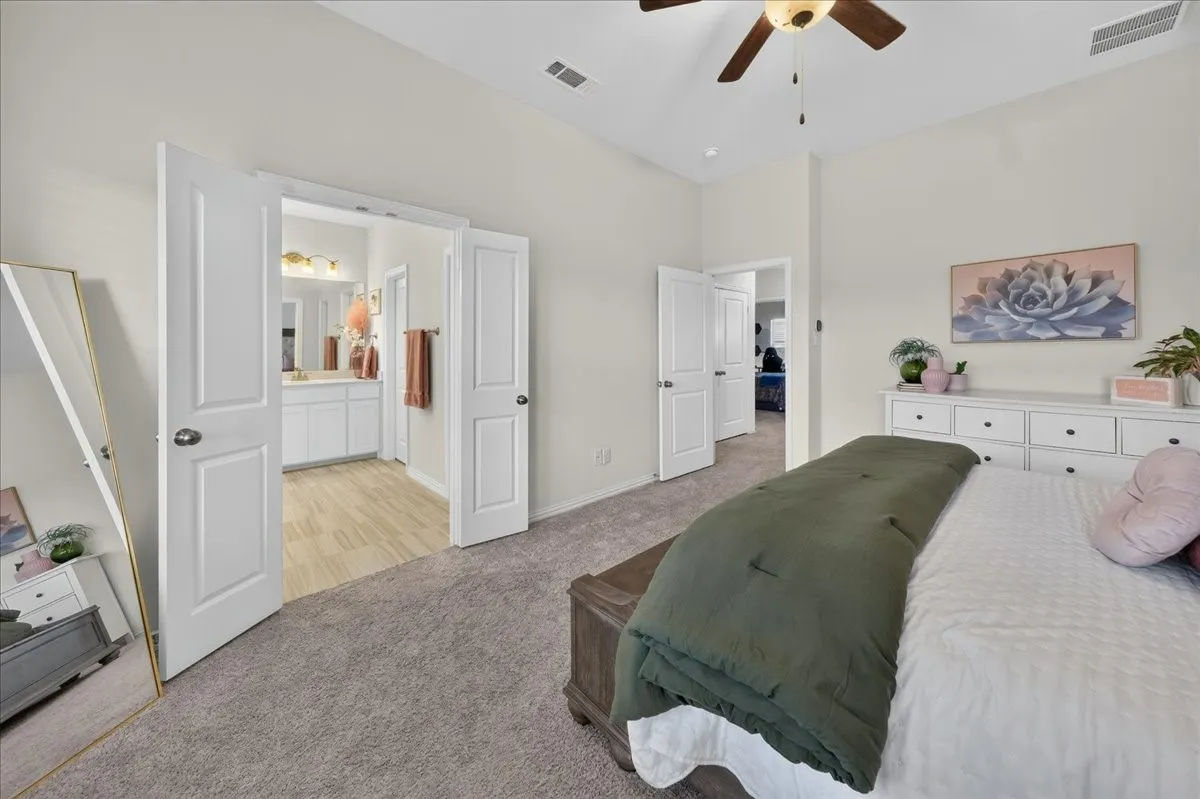 Vaulted ceiling bedroom with soft gray carpeting, featuring an en-suite bathroom with light wood-finish flooring and a double vanity