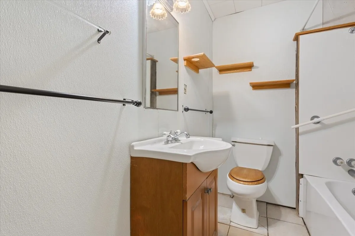 Bathroom featuring a wood-finish vanity with an integrated sink, a commode with a wood-finish seat, a bathtub, and wall-mounted shelving