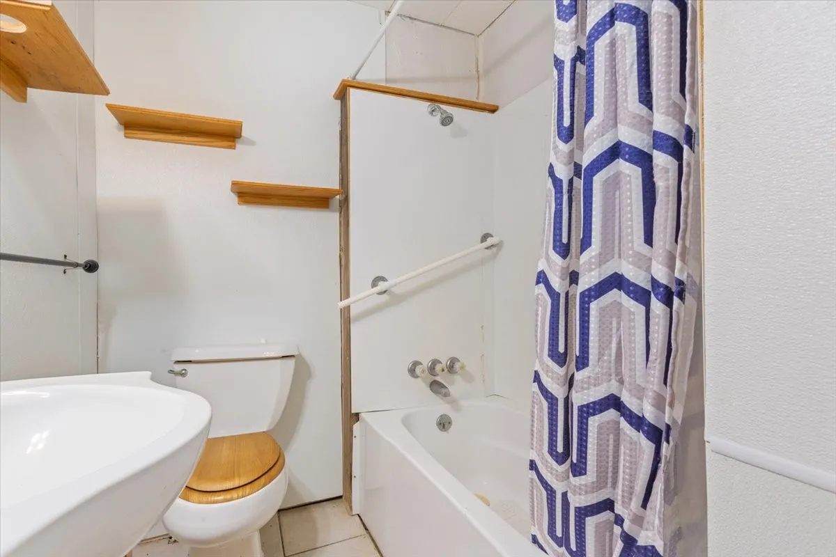 Bathroom featuring a white porcelain sink, toilet with wood-finish seat, built-in tub with shower, and decorative wall shelving