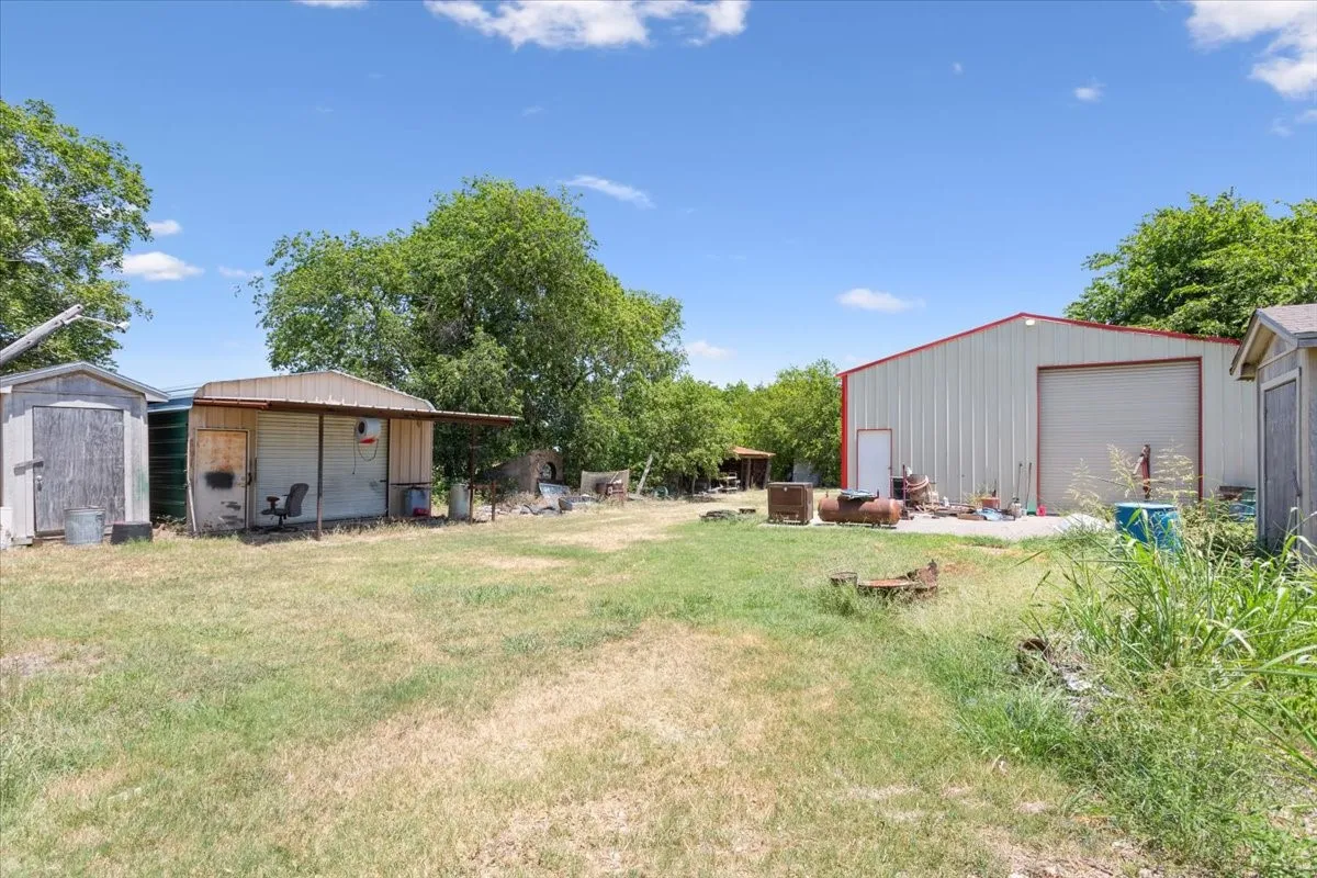 Expansive open-air lot featuring a large metal building with an overhead door, multiple storage sheds, and mature perimeter trees