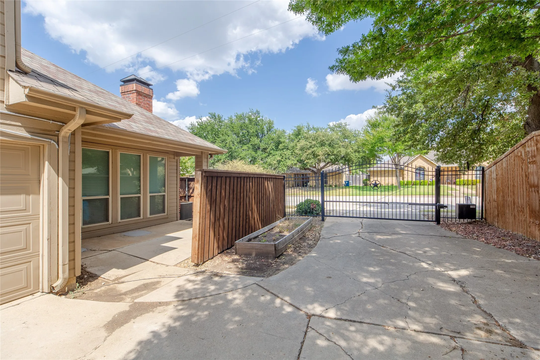 Gated driveway access featuring a wrought iron gate, concrete surfacing, and surrounding wood fencing