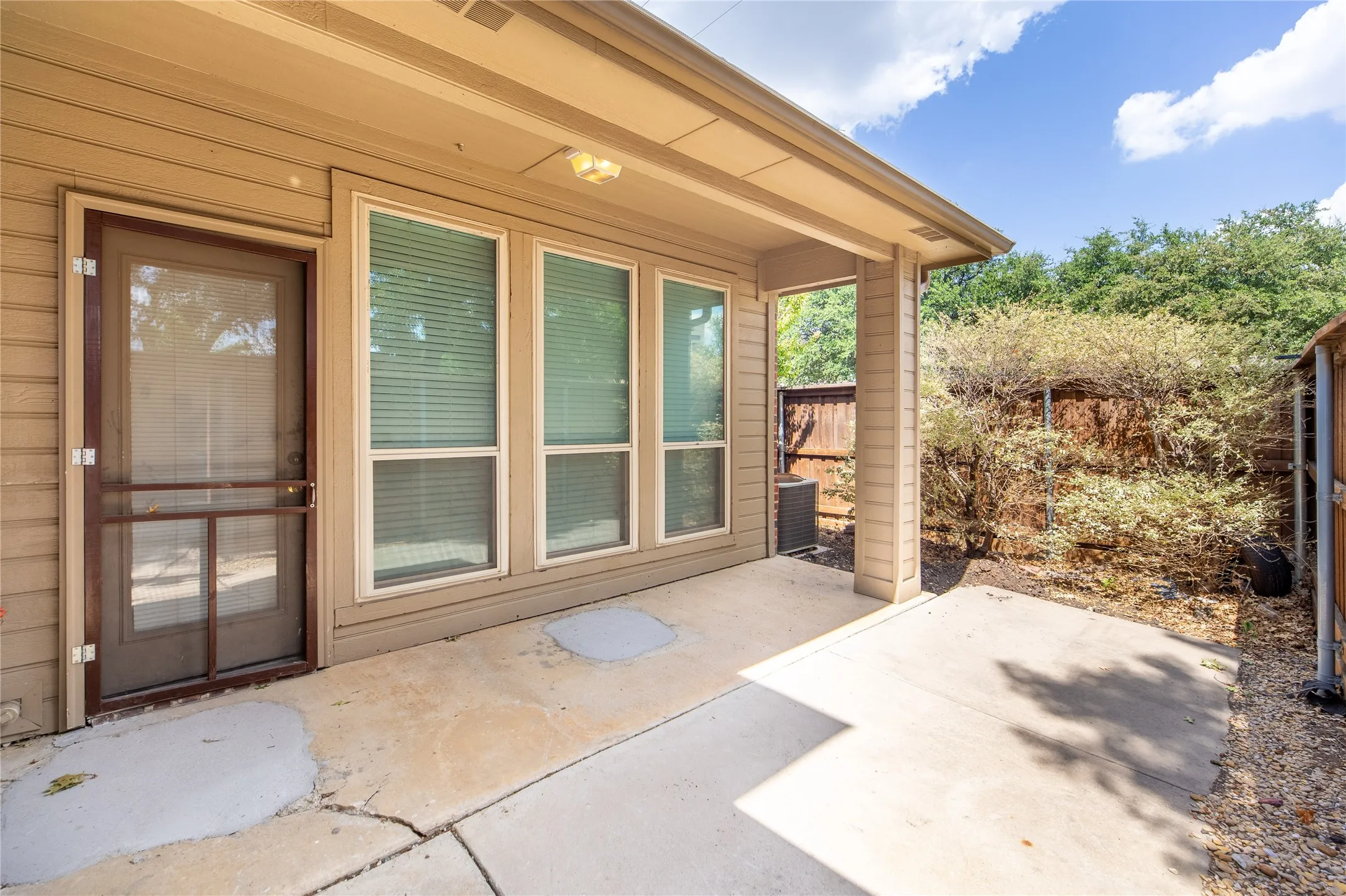 Patio space featuring concrete flooring, a covered overhang, three large windows with blinds, a screen door, and a privacy fence