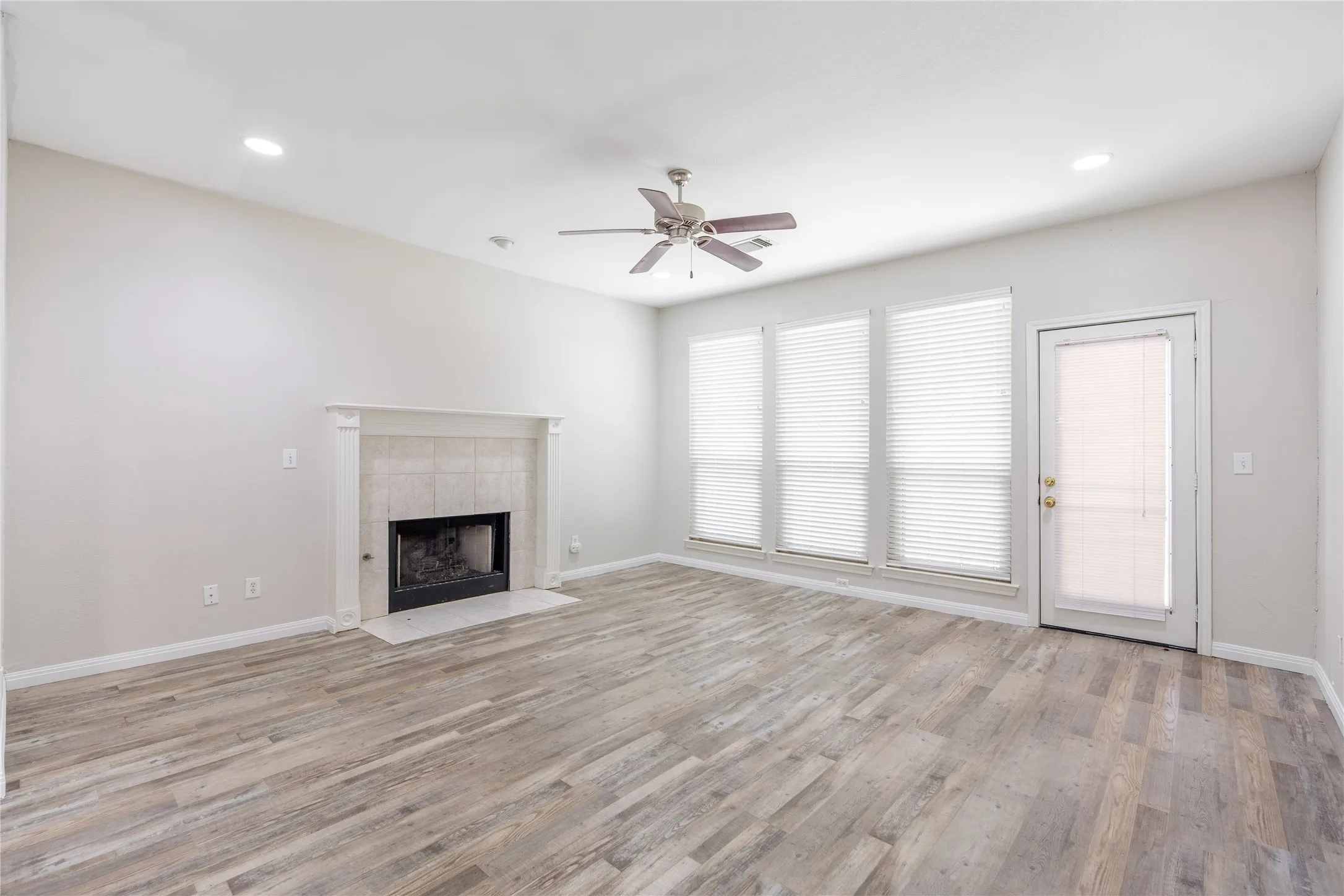 Spacious room featuring wood-finish flooring, a tile-surround fireplace with a classic mantel, and multiple windows with blinds