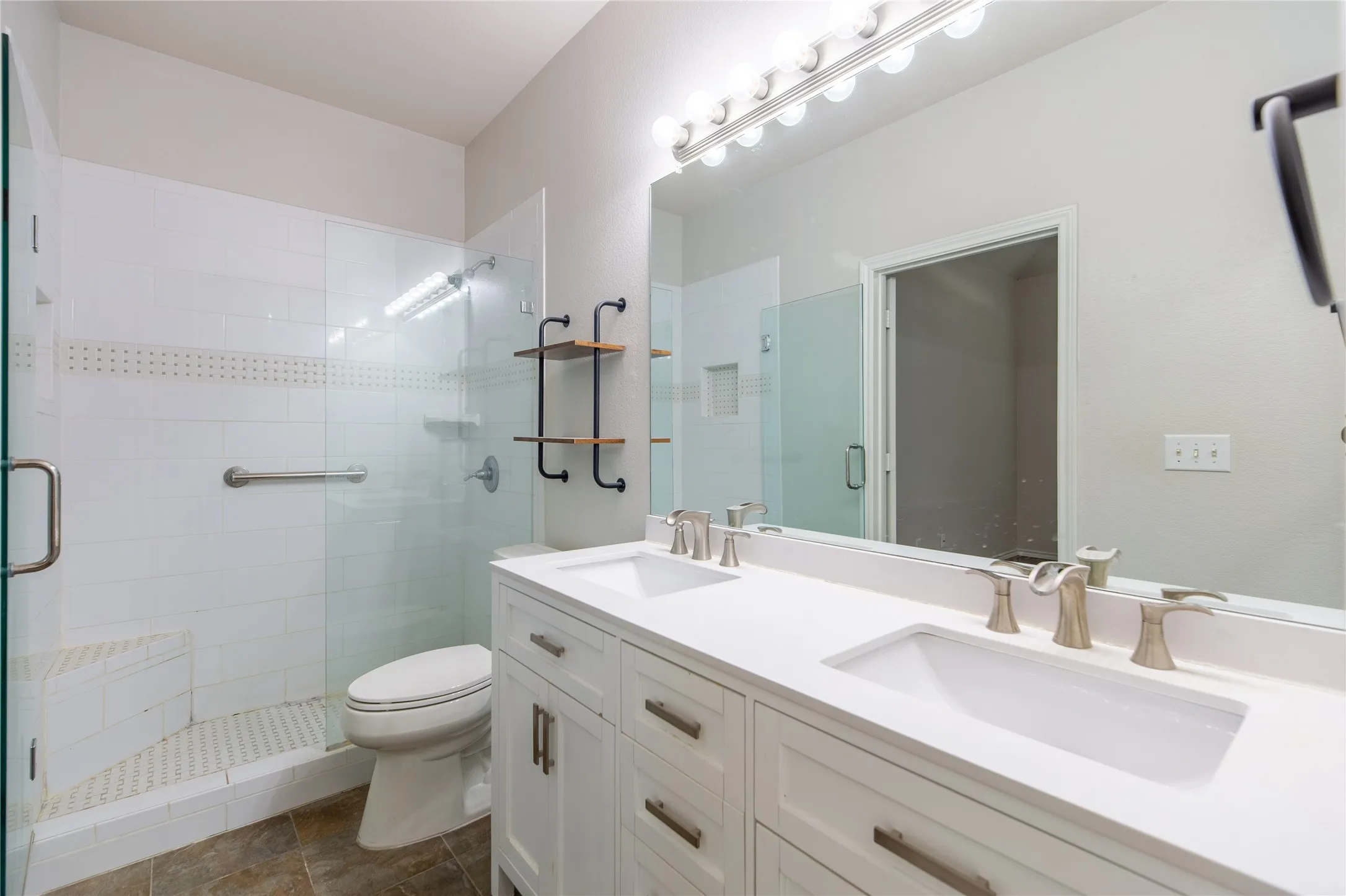 Modern bathroom featuring a dual vanity with white cabinetry and integrated sinks, brushed nickel fixtures, a large wall-mounted mirror with overhead lighting, and a walk-in shower with clear glass enclosure and white tiled walls