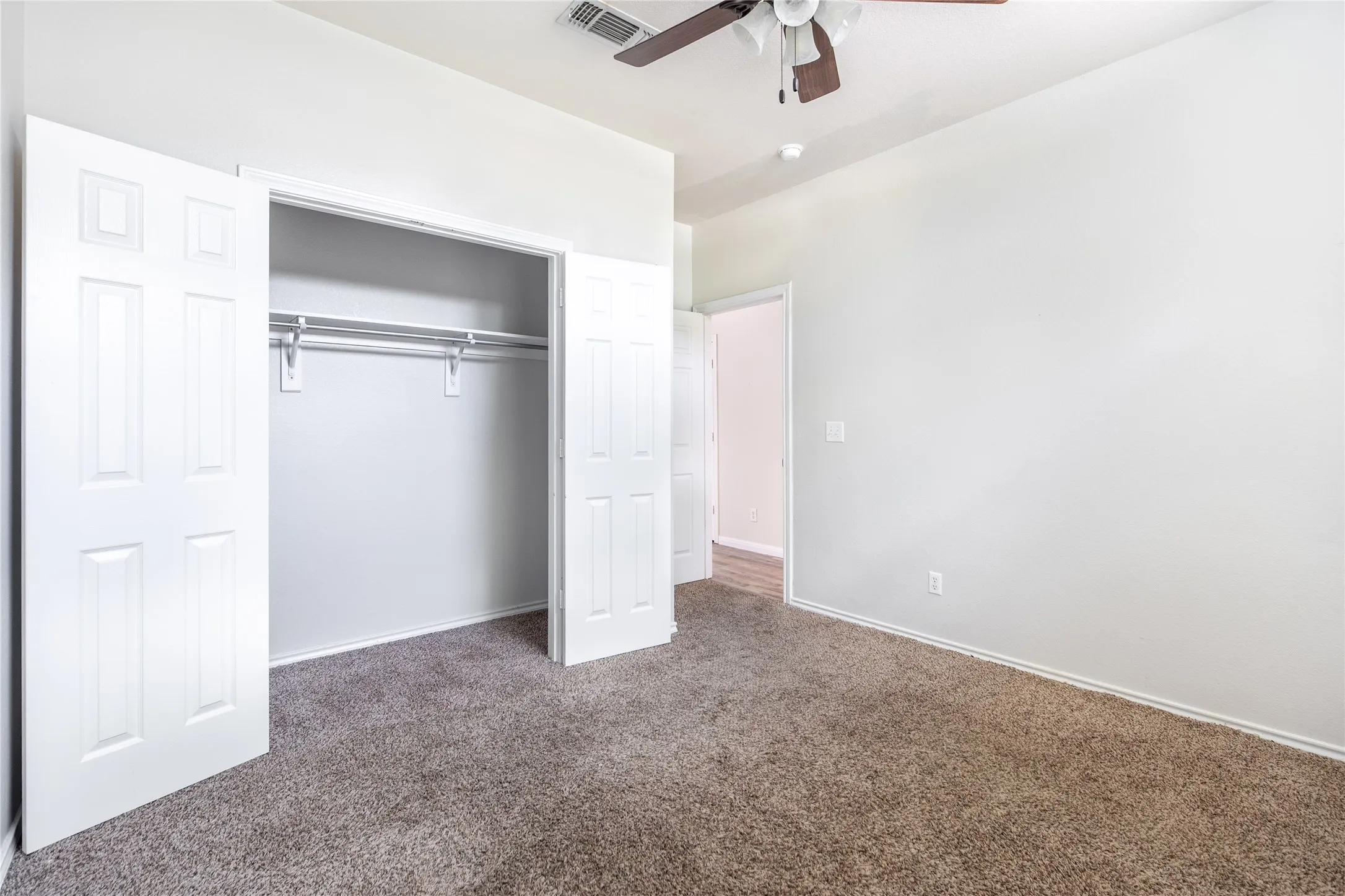Neutral-toned carpeted room featuring a ceiling fan with integrated lighting, a spacious closet with bi-fold panel doors and dual clothing rods, and white painted walls