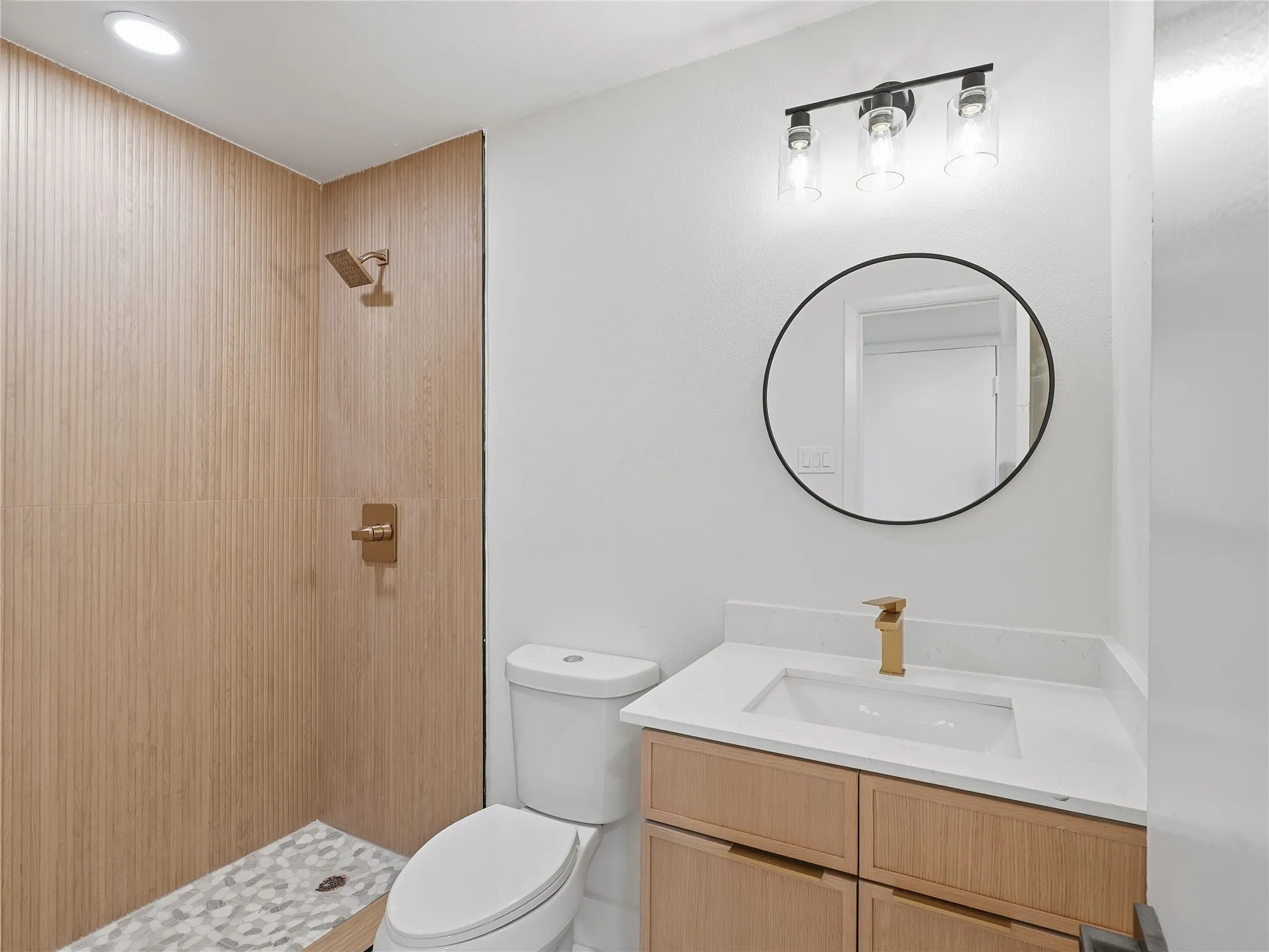Modern bathroom featuring a walk-in shower with wood-finish wall tiles and a patterned floor, a light wood vanity with a white countertop and integrated sink, a circular mirror, and contemporary black and brass fixtures