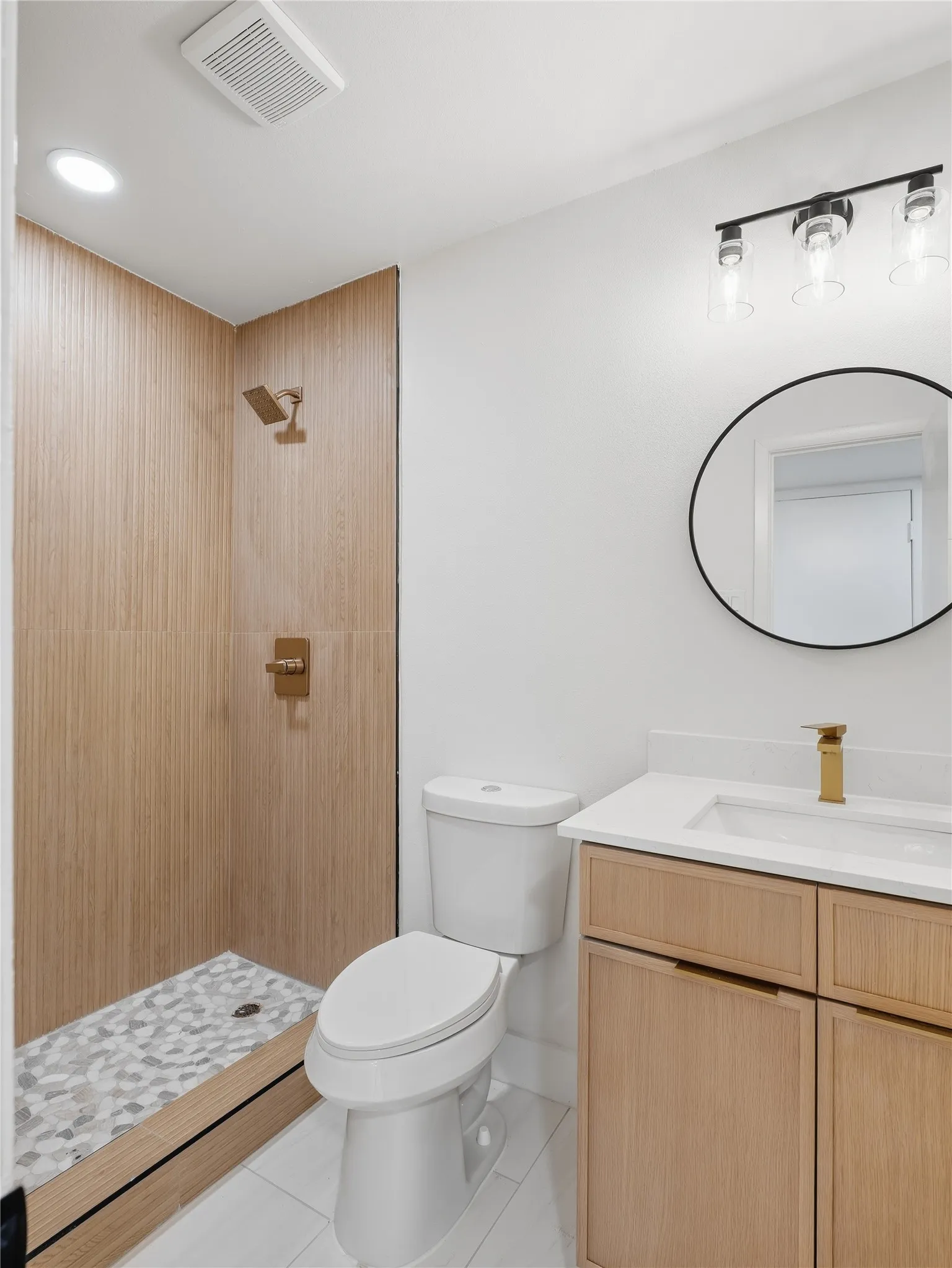 Bathroom featuring a walk-in shower with wood-finish tile and pebble flooring, white floor tile, a light wood-finish vanity with a white countertop and gold-tone faucet, and a round black-framed mirror
