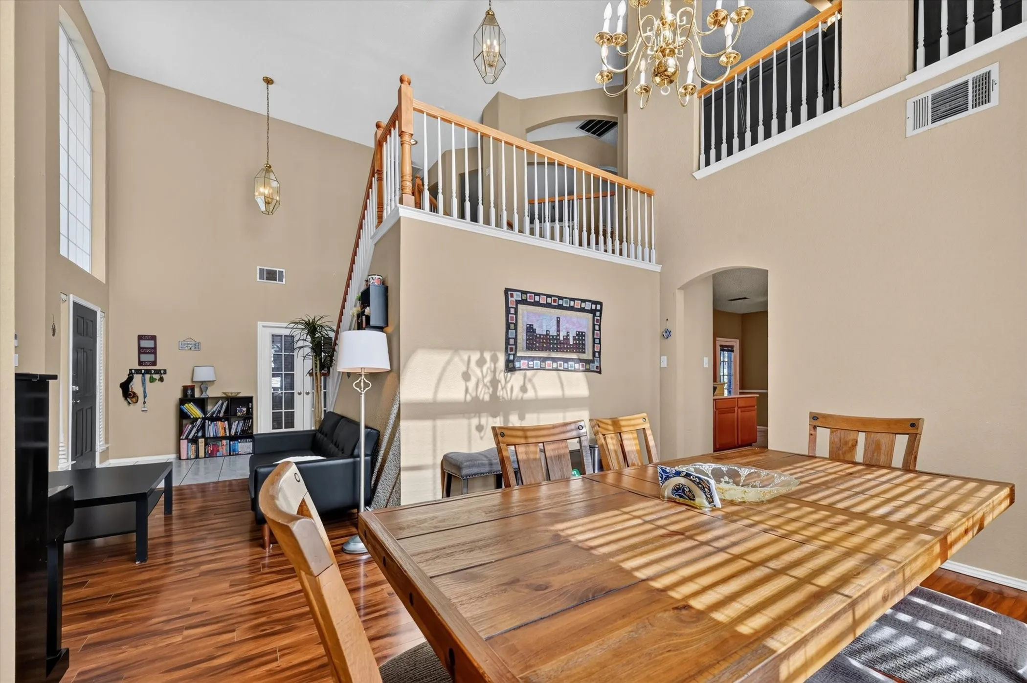 Spacious living area featuring double-height ceilings, wood-finish flooring, an upper-level balcony, and a prominent dining area with a chandelier