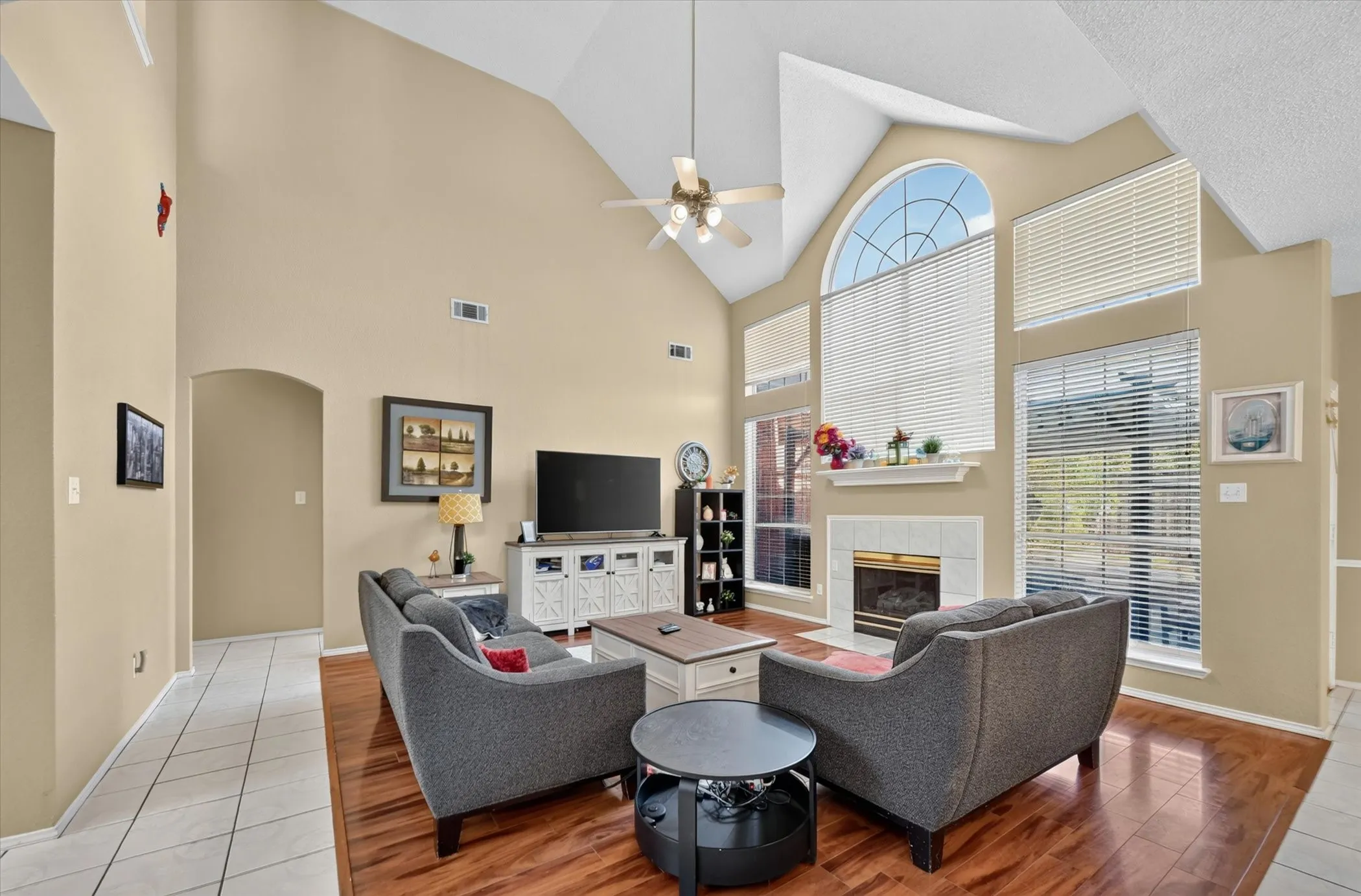Vaulted ceiling living area featuring a fireplace with a tile surround, large windows, wood-finish flooring, and a ceiling fan