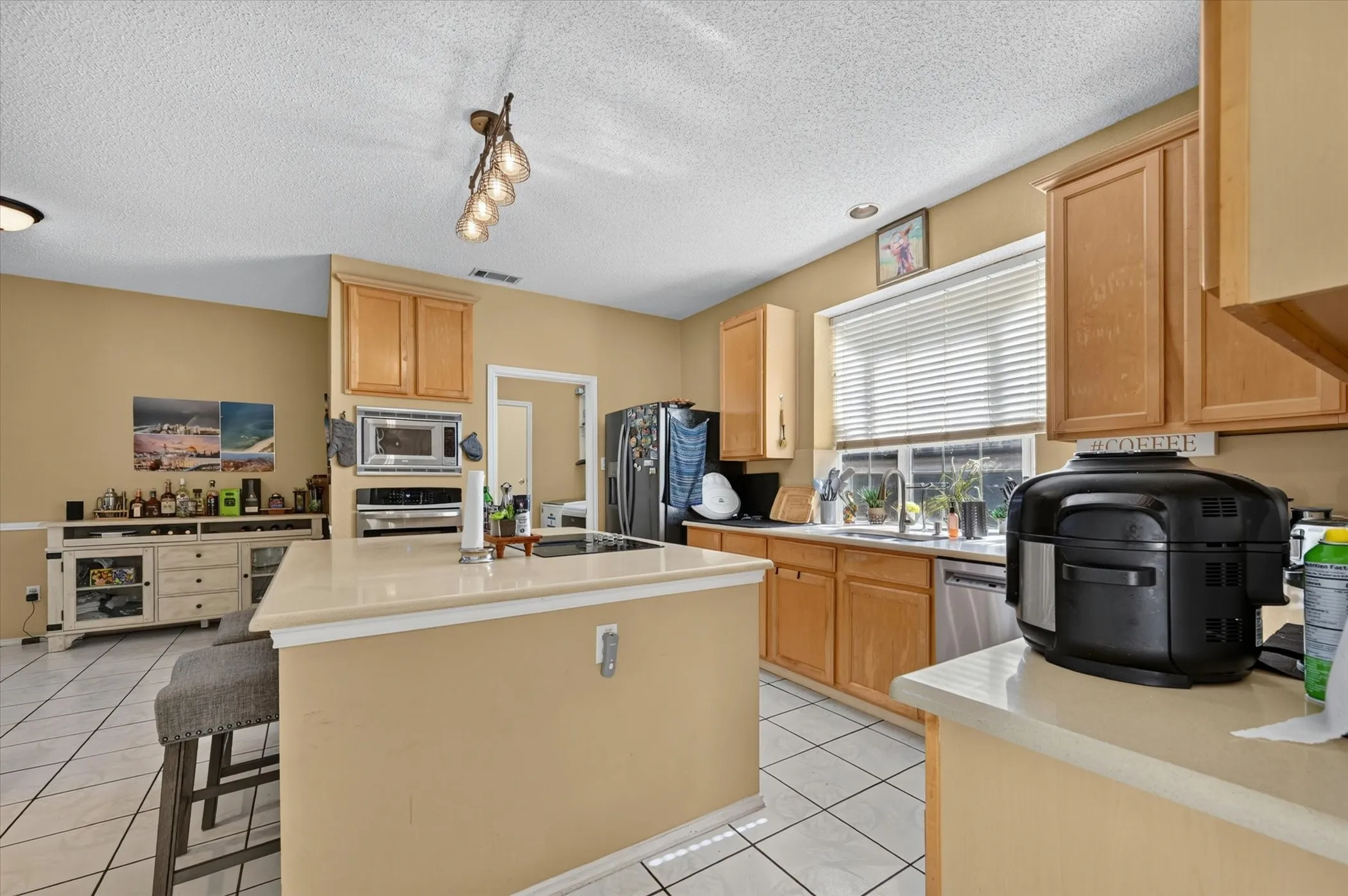 Kitchen featuring light wood cabinetry, white tile flooring, a central island with a light-toned countertop, stainless steel appliances, and a window with horizontal blinds