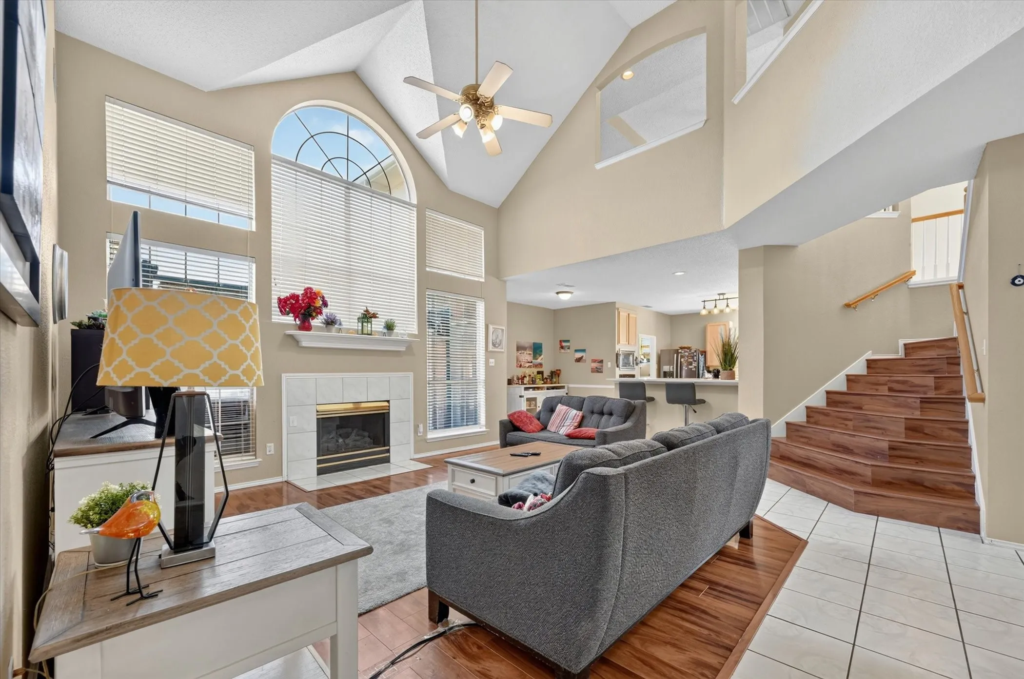 Two-story living area featuring a vaulted ceiling with a ceiling fan, a fireplace with a white tile surround and mantel, and large windows with an arched top window