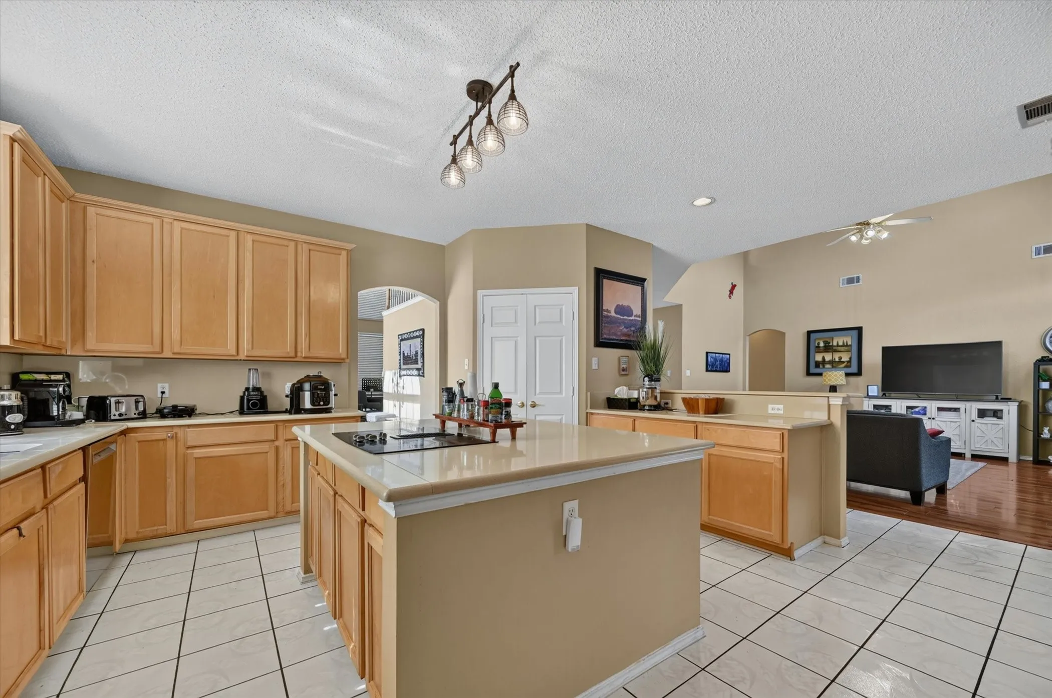 Spacious kitchen featuring light wood cabinetry, light-toned countertops, a central island with a cooktop, and tile flooring
