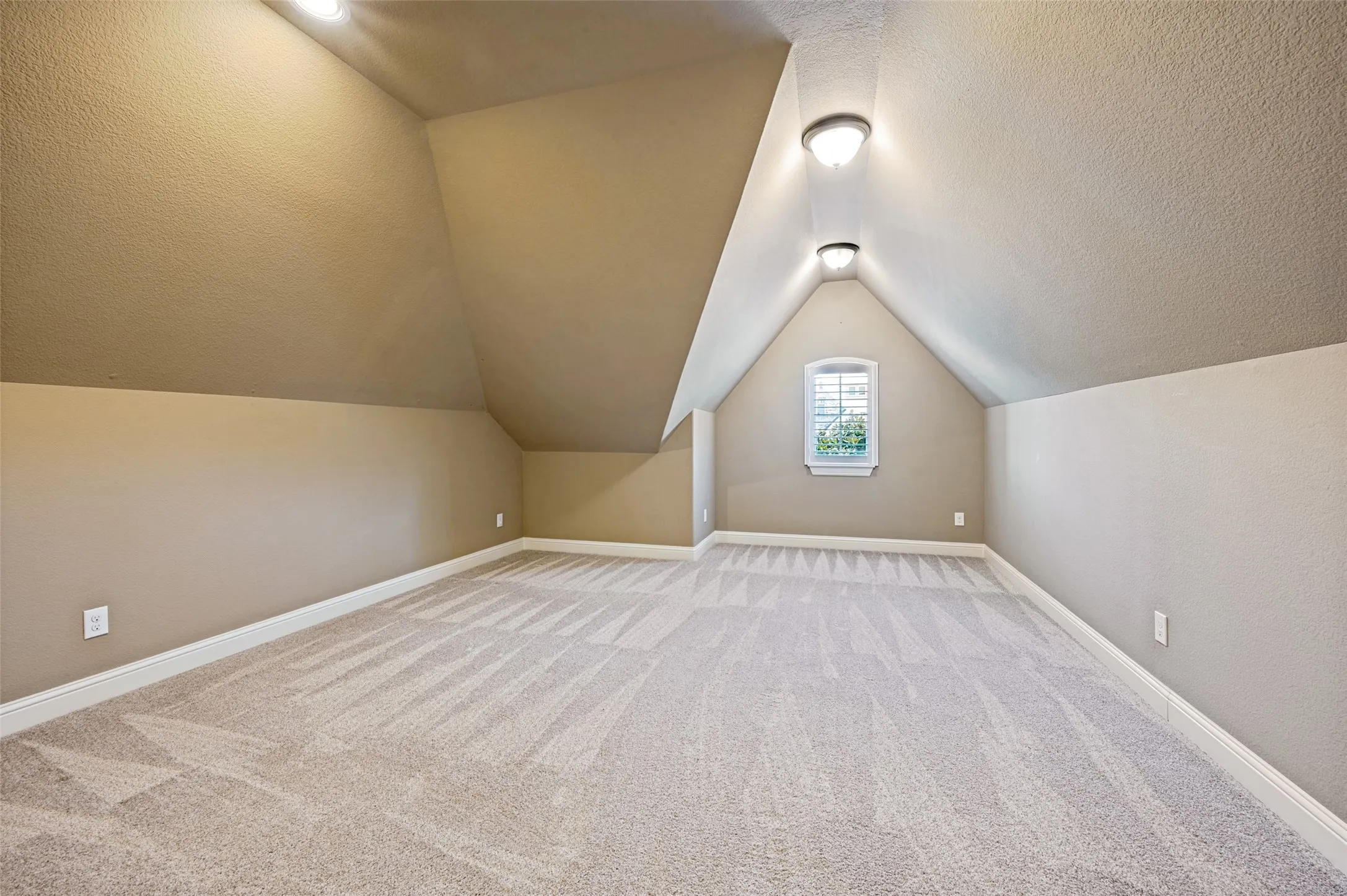 Spacious room featuring vaulted ceilings, a single window with plantation shutters, neutral-toned carpeting, and a pair of semi-flush mount ceiling lights