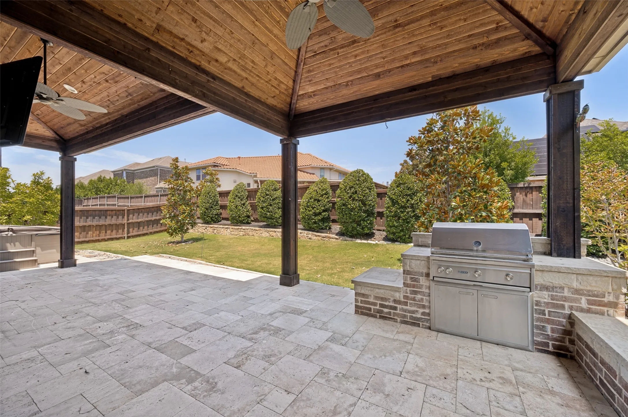 Covered outdoor patio featuring stone-look pavers, a built-in stainless steel grill, a wood-finish ceiling with integrated ceiling fans, and a spacious grassy yard