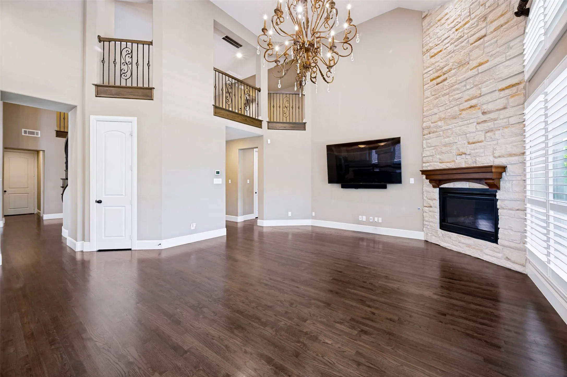 Spacious living area featuring high ceilings, wood-finish flooring, a stone fireplace with a wood mantel, a decorative chandelier, and interior balconies with wrought iron railings