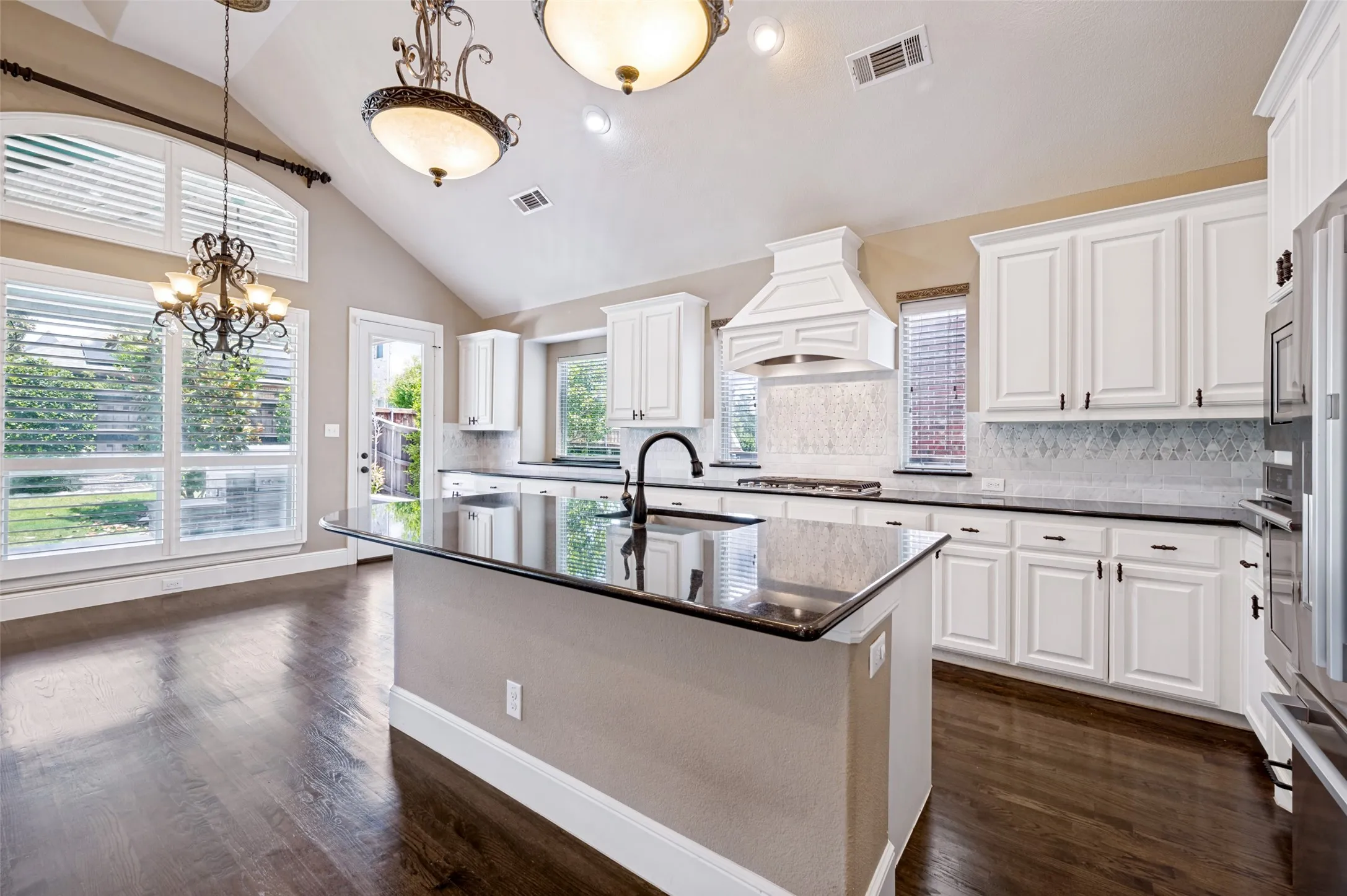 Kitchen featuring dark wood-finish flooring, a central island with a dark countertop, white cabinetry with dark hardware, a decorative range hood, and a light-toned tile backsplash