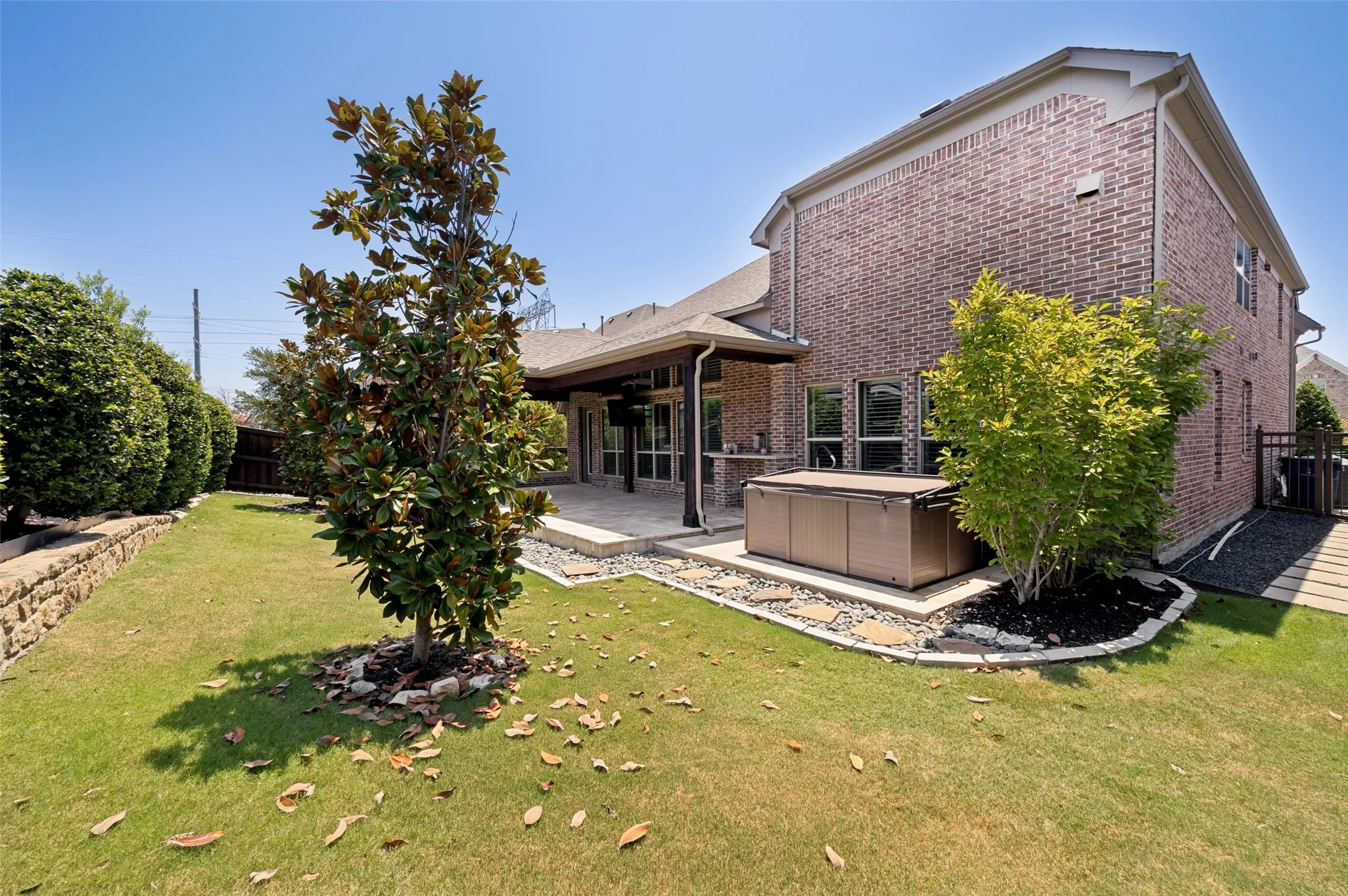 Expansive backyard featuring a covered patio, brick exterior, lush green lawn, mature trees, and a stone retaining wall
