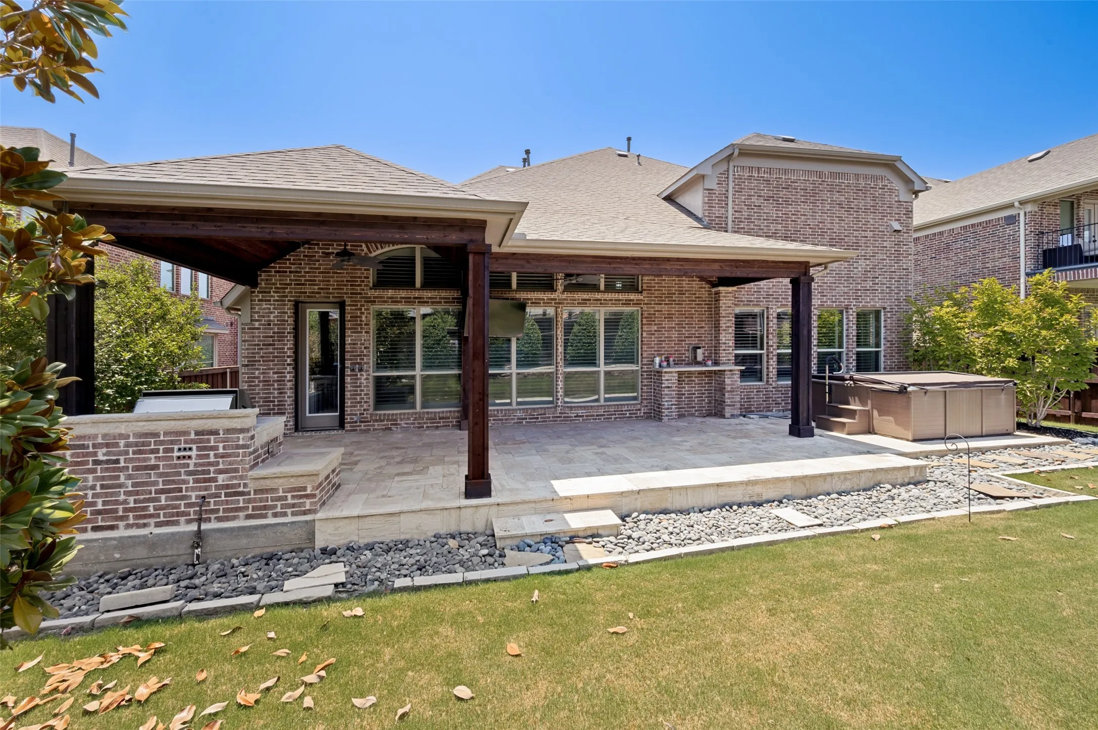 Expansive outdoor living area featuring a covered patio with dark wood beams, brick exterior, built-in outdoor kitchen, hot tub, and stone rock landscaping