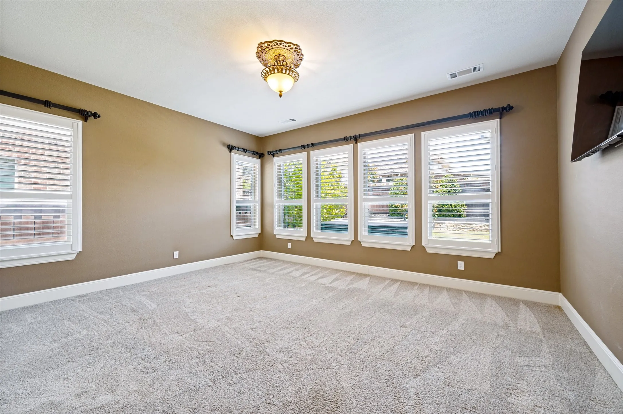Expansive room featuring light gray patterned carpeting, a decorative ceiling light fixture, multiple windows with white plantation shutters, and warm neutral wall paint
