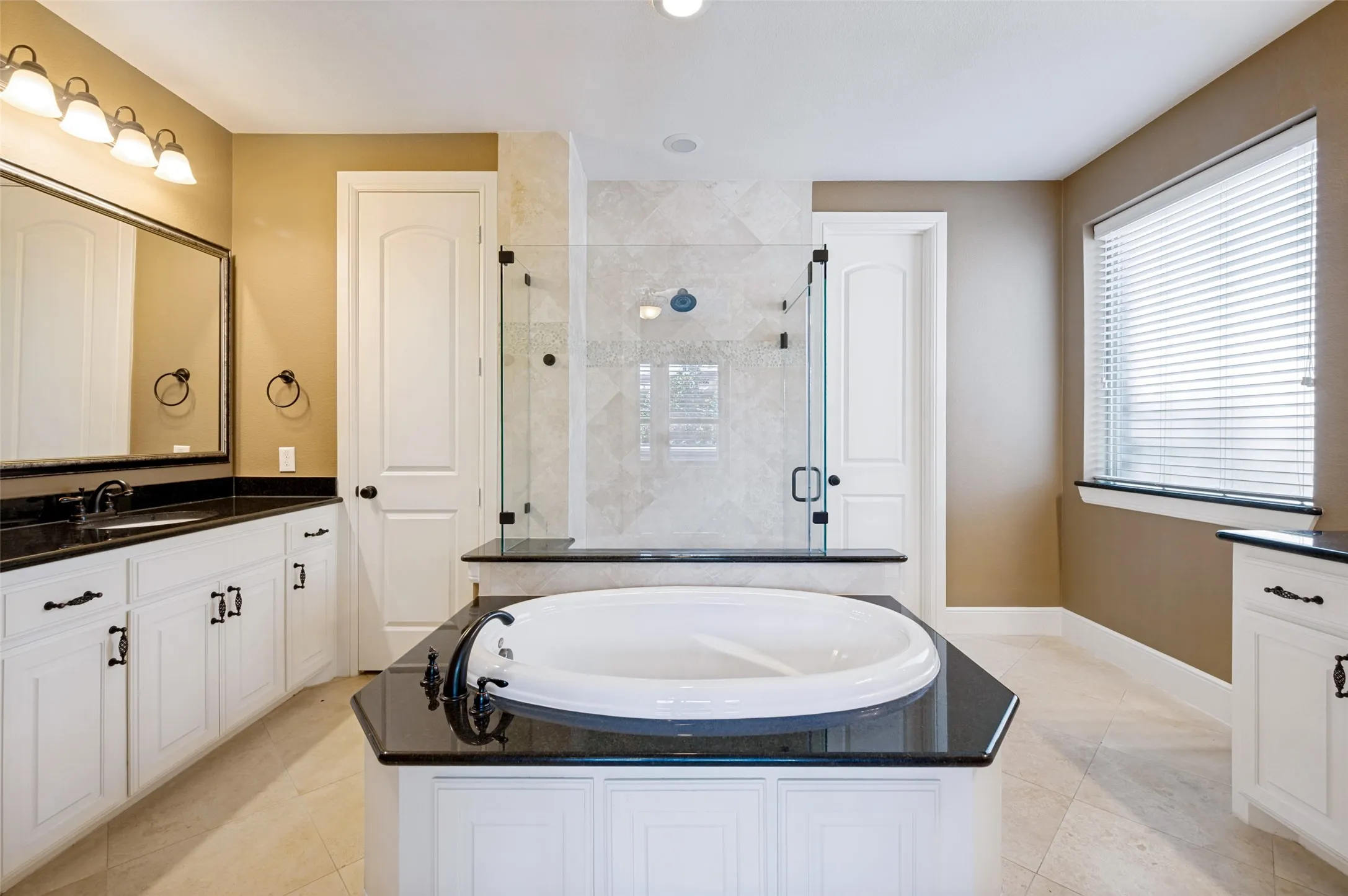 Bathroom featuring a large oval soaking tub with a black countertop surround, a walk-in shower with clear glass enclosure and tiled walls, dual vanities with white cabinetry and dark countertops, and tile flooring