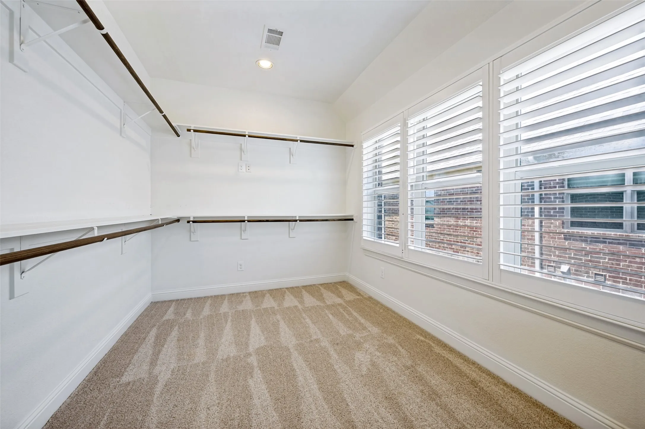 Spacious closet featuring extensive shelving and hanging rods, patterned carpet, and three windows with white plantation shutters