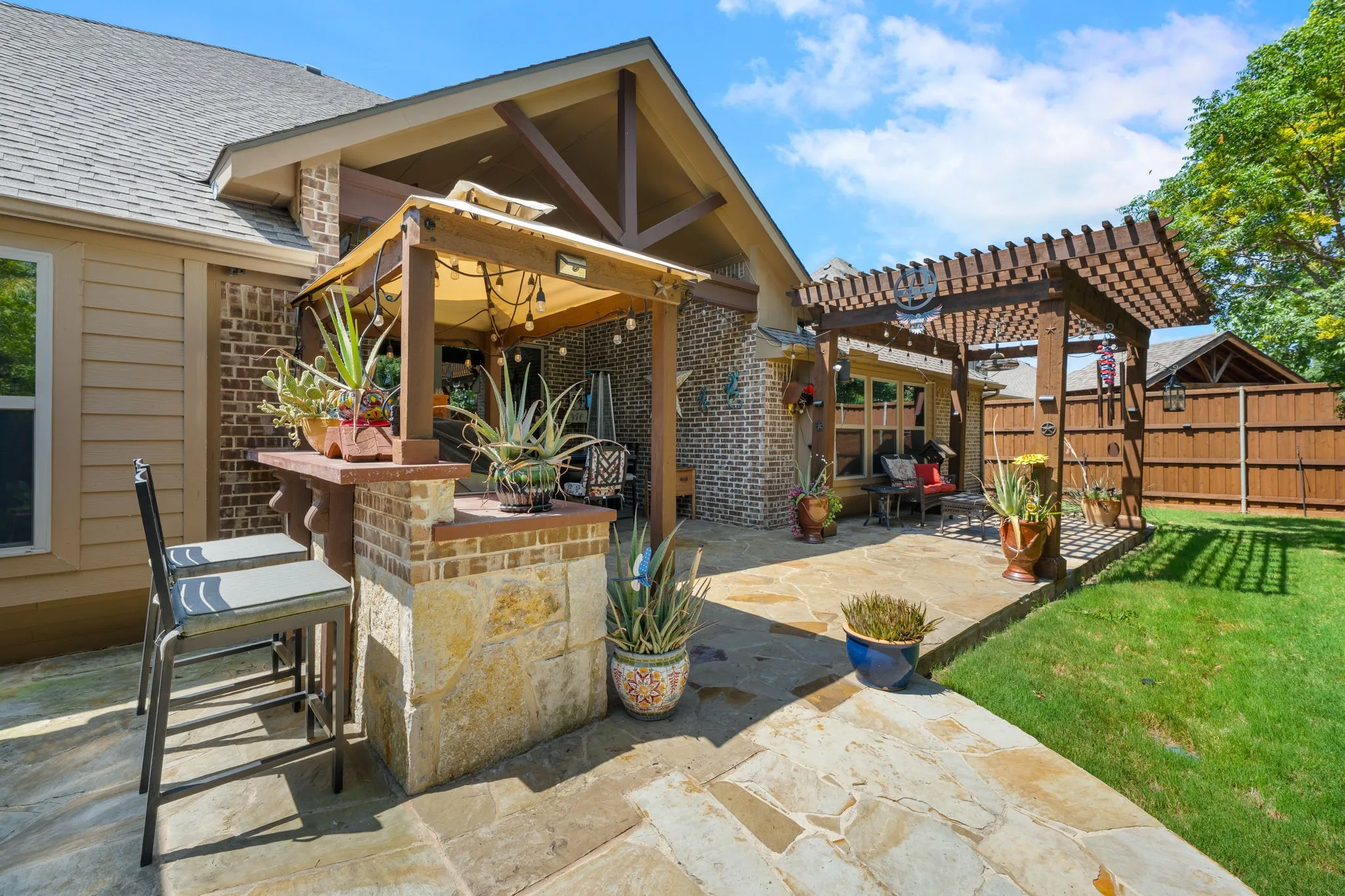 Expansive flagstone patio featuring a stone and brick outdoor bar, a covered outdoor living area with exposed beams, a wood pergola, and a manicured lawn