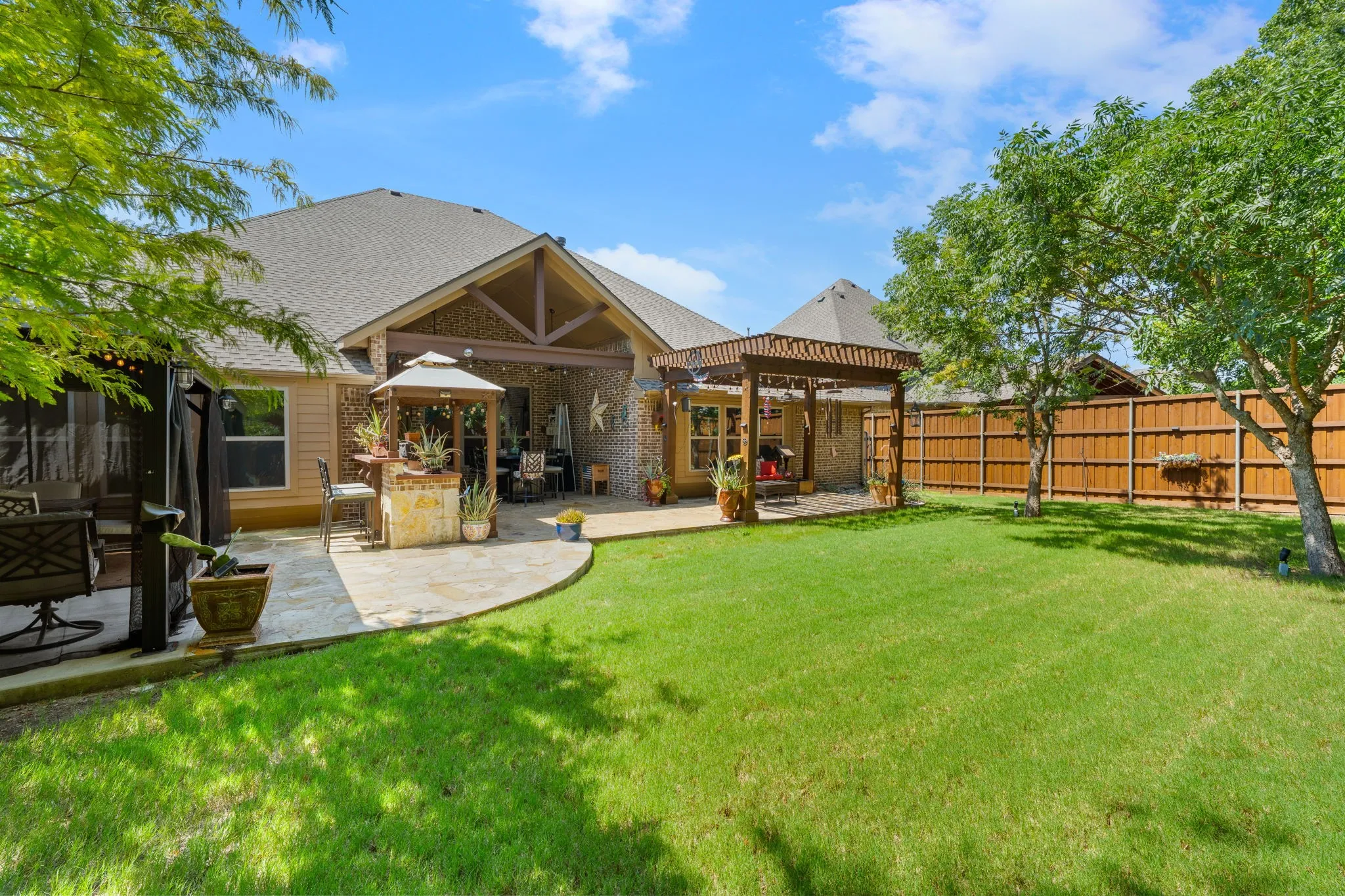 Expansive backyard featuring a stone patio, covered porch with exposed beams, wood pergola, and perimeter wood fencing