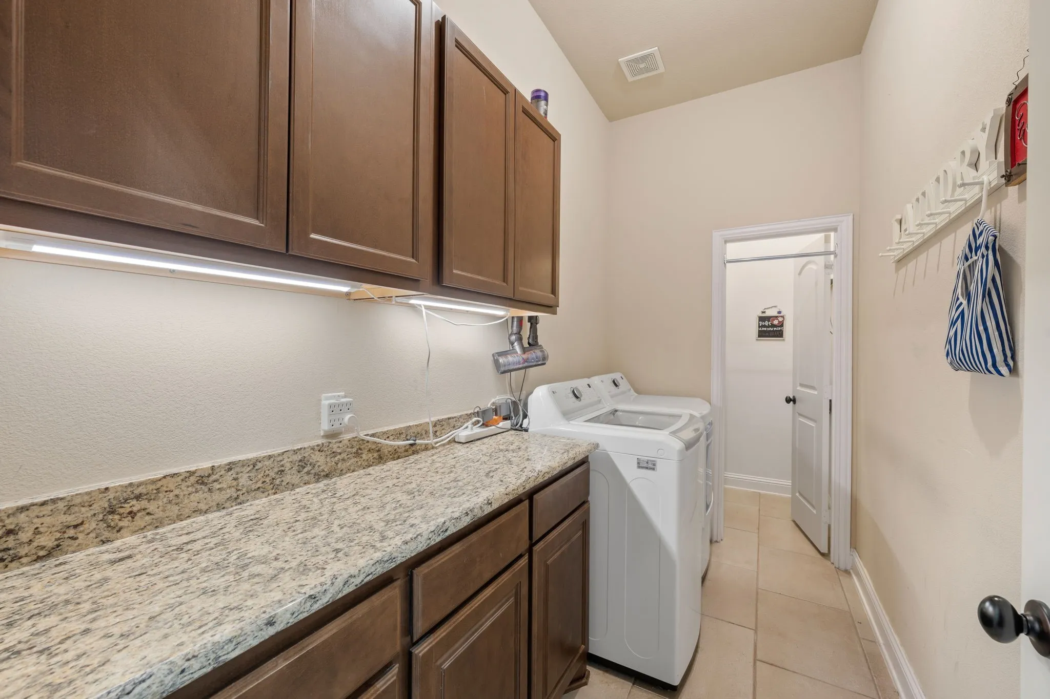 Look at the size of this laundry room! Laundry room featuring granite-look countertops, wood-finish cabinetry, under-cabinet lighting, and tiled flooring