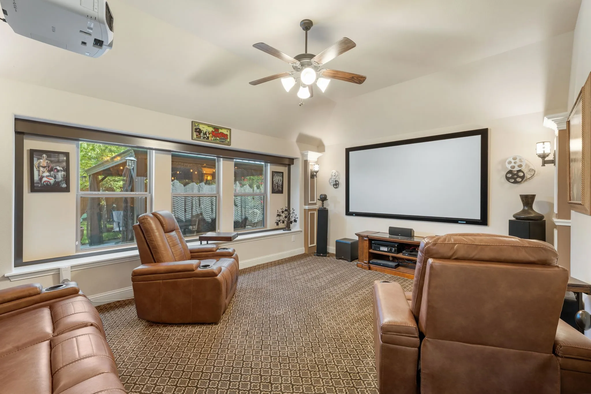 Dedicated media room featuring a large projection screen, vaulted ceiling with a wood-finish ceiling fan, patterned carpet flooring, extensive window seating, and built-in wall sconces