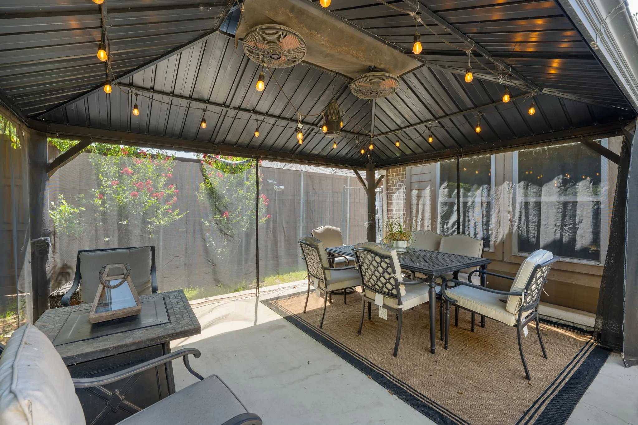 Covered outdoor patio featuring a metal gazebo with string lighting, dual ceiling fans, and clear screen walls