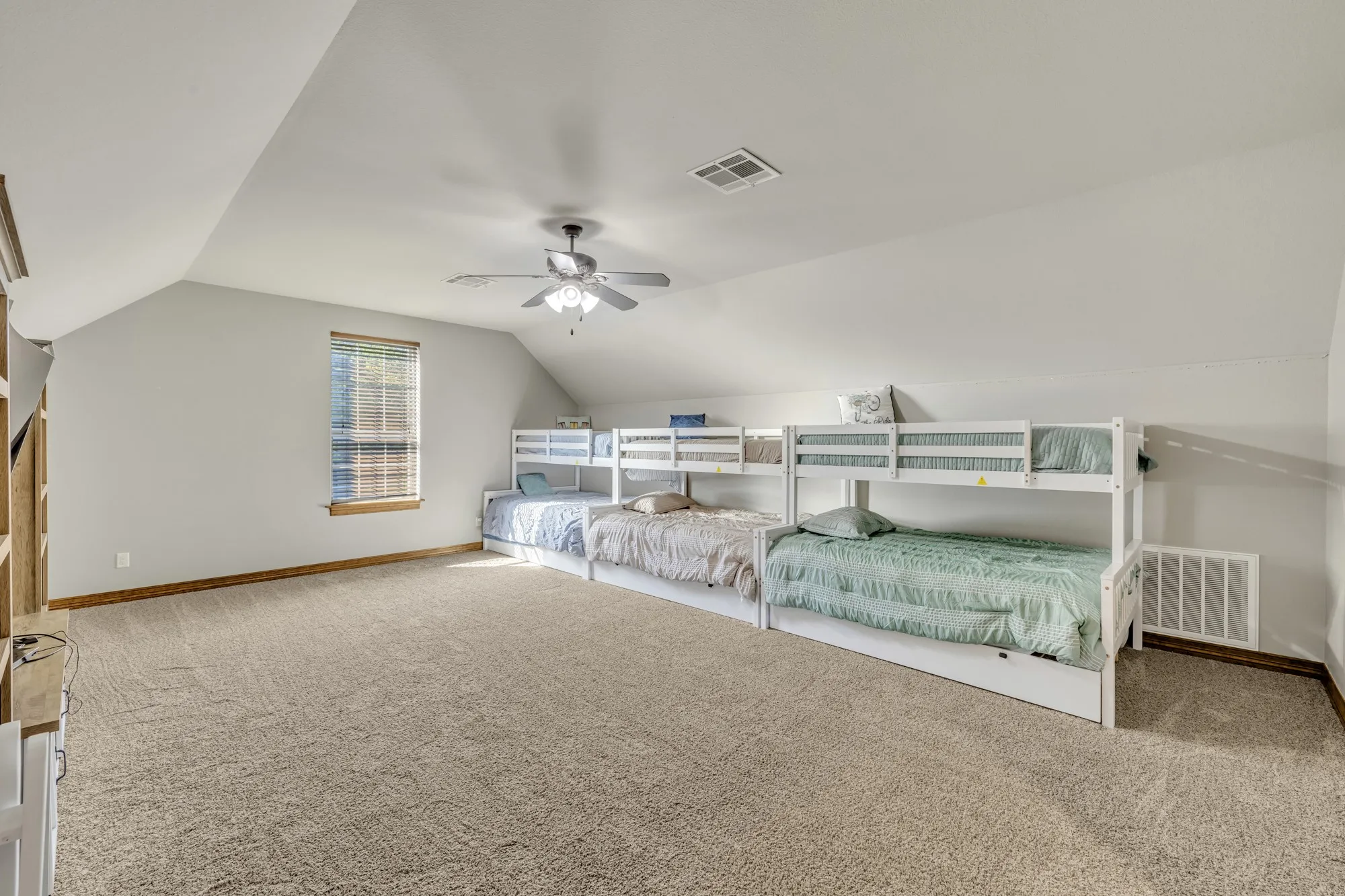 Spacious room featuring sloped ceilings, plush carpeting, a ceiling fan with integrated lighting, window with blinds, and built-in shelving
