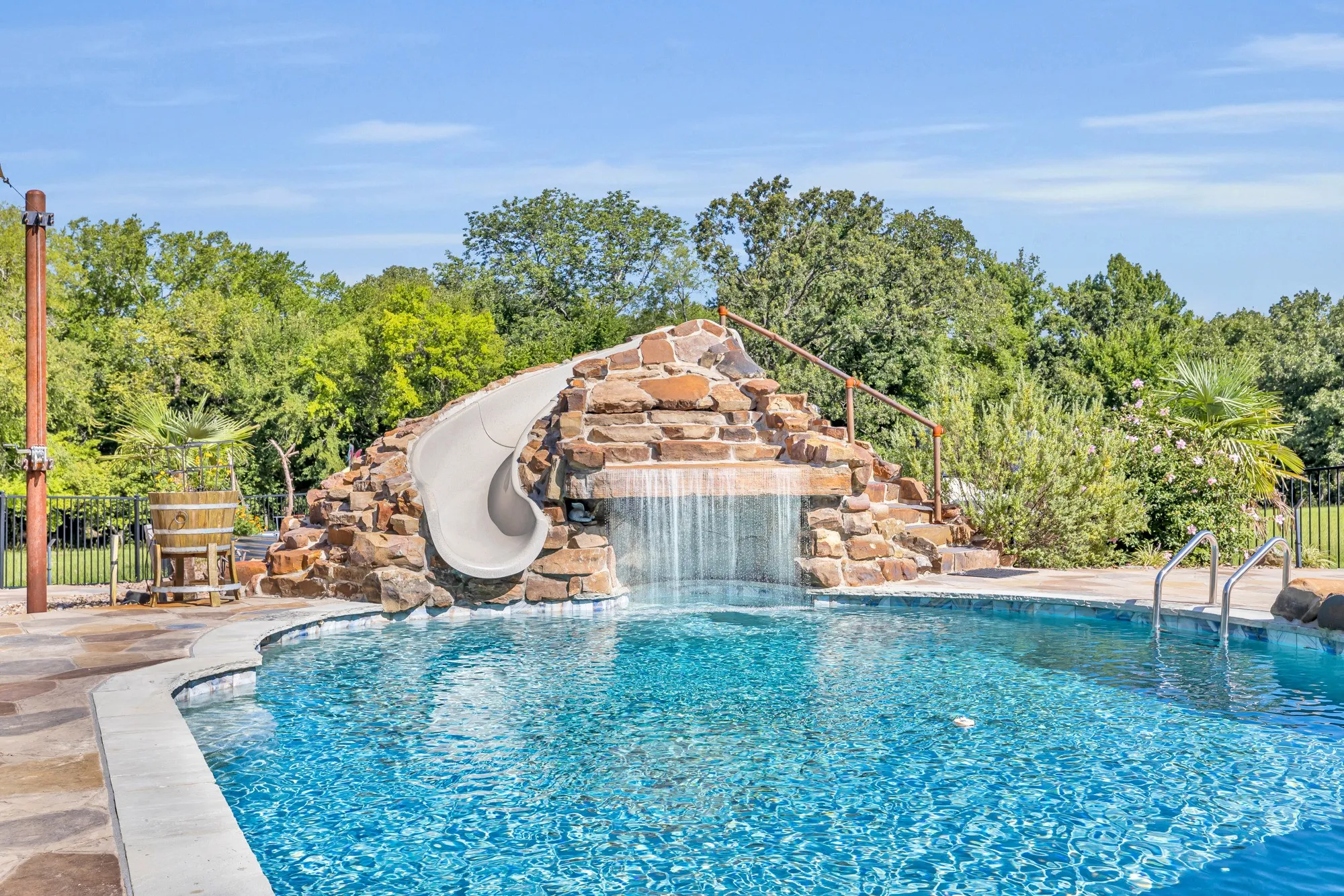 Curved swimming pool featuring a rock waterfall, built-in waterslide, stone coping, and flagstone deck