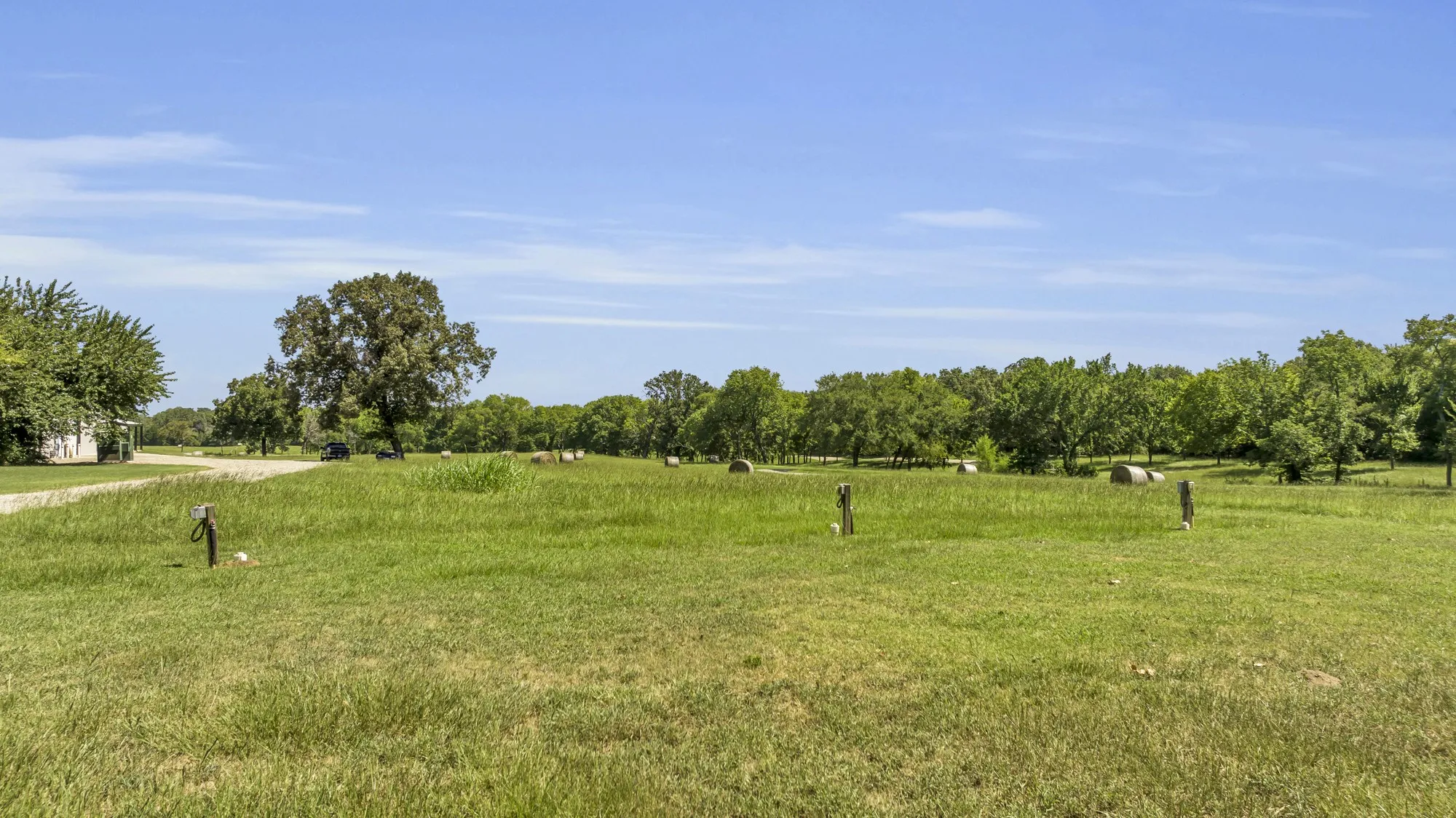 Expansive green field with mature trees, scattered hay bales, and a clear blue sky