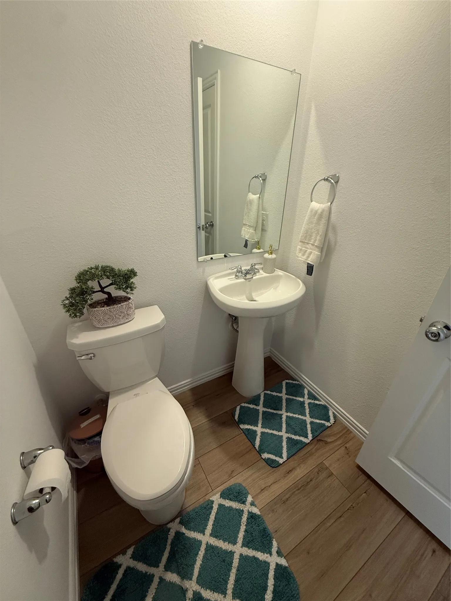 Pedestal sink with chrome fixtures, wood-finish flooring, wall-mounted mirror, and a white toilet