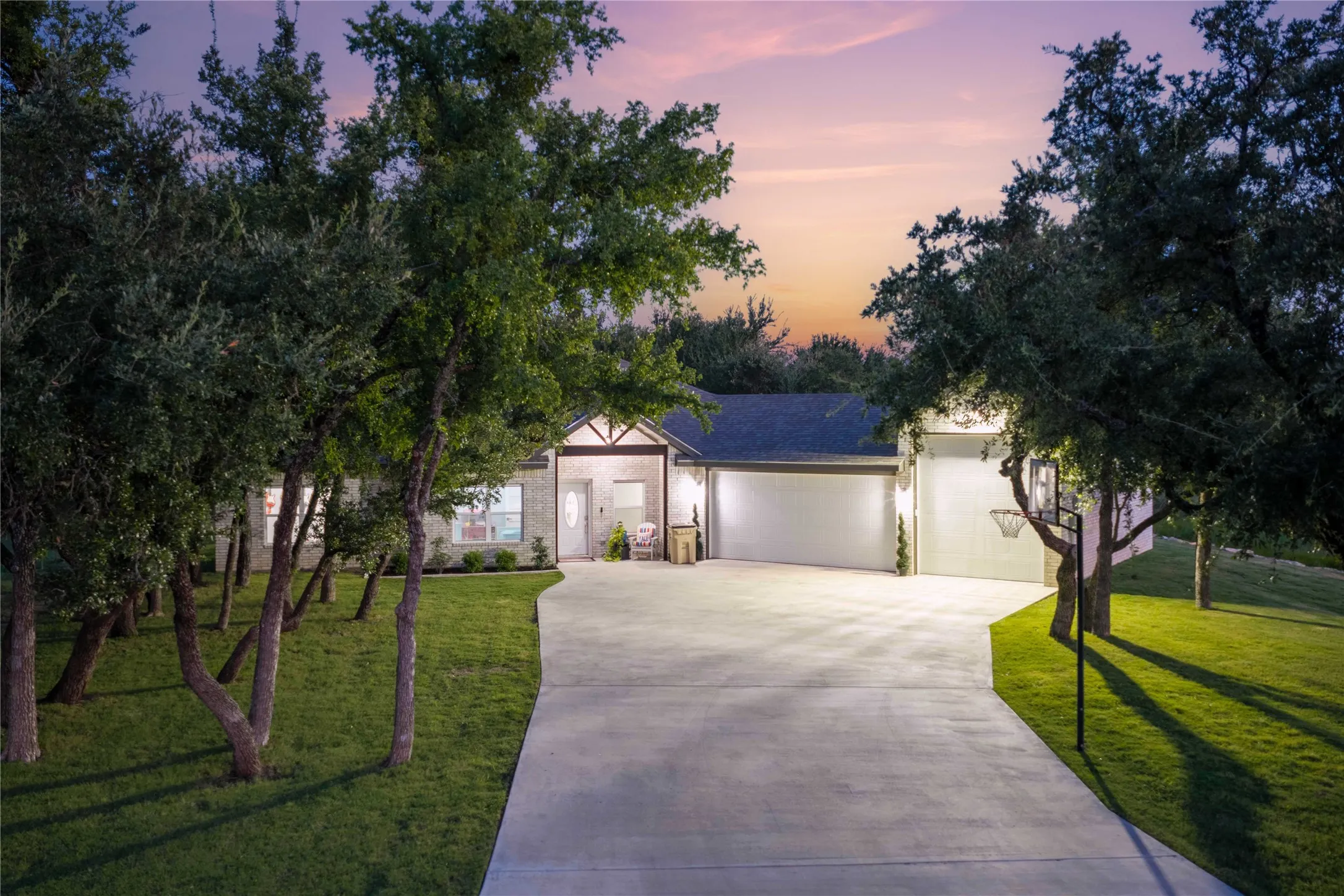 Stone facade residence featuring a prominent two-car garage, mature trees, a concrete driveway, and a manicured lawn