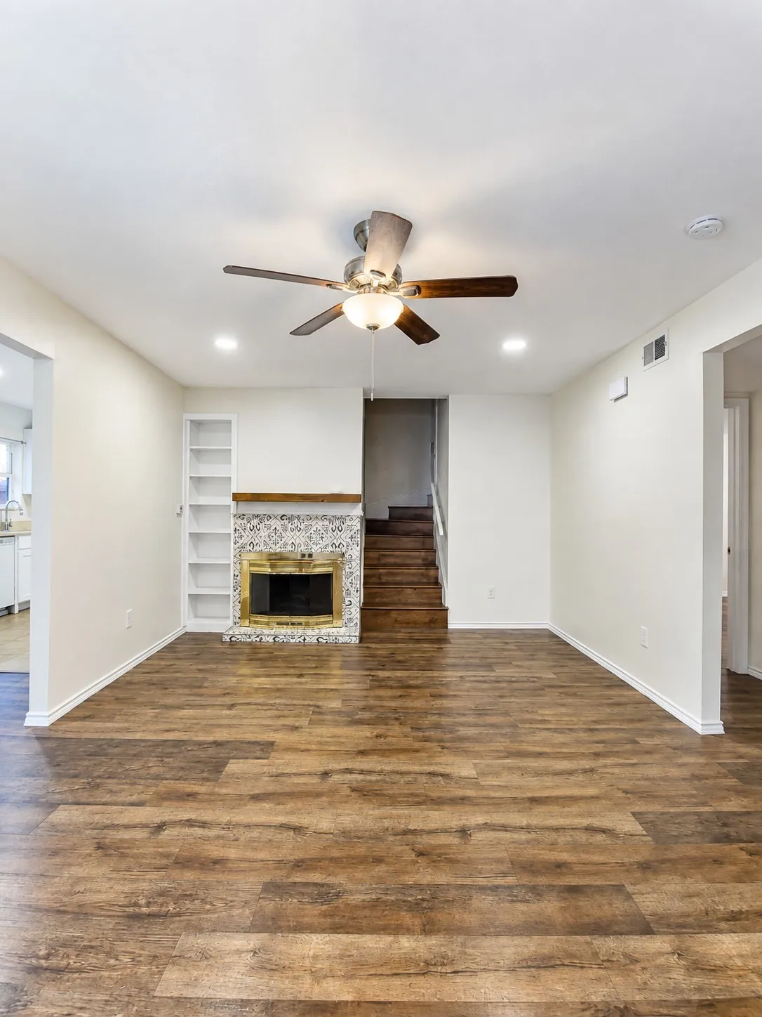 Wood-finish flooring throughout, fireplace with decorative tile surround and brass-finish insert, built-in shelving, and a ceiling fan with integrated lighting