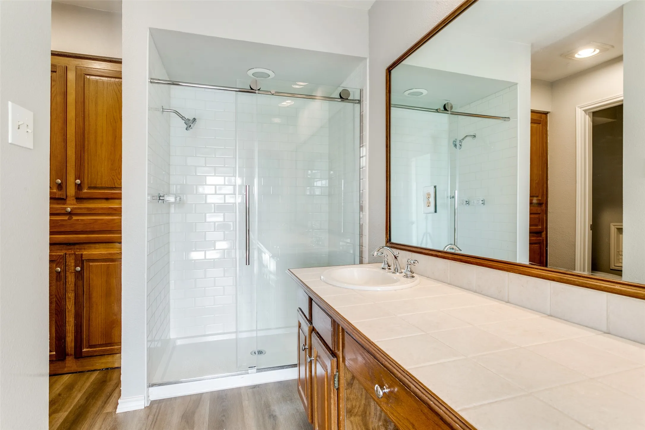 Bathroom featuring a glass-enclosed shower with subway tile, wood-finish flooring, a wood vanity with a tile countertop, and a large wall-mounted mirror