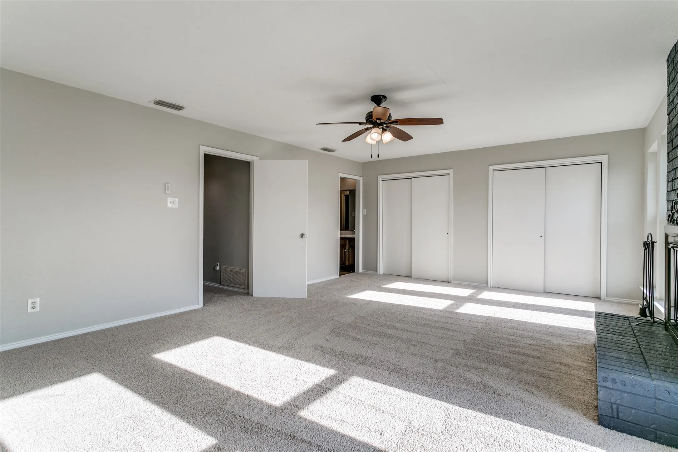 Spacious room featuring light gray carpeting, a ceiling fan with integrated lighting, two sets of sliding closet doors, and a brick fireplace with an iron fireguard