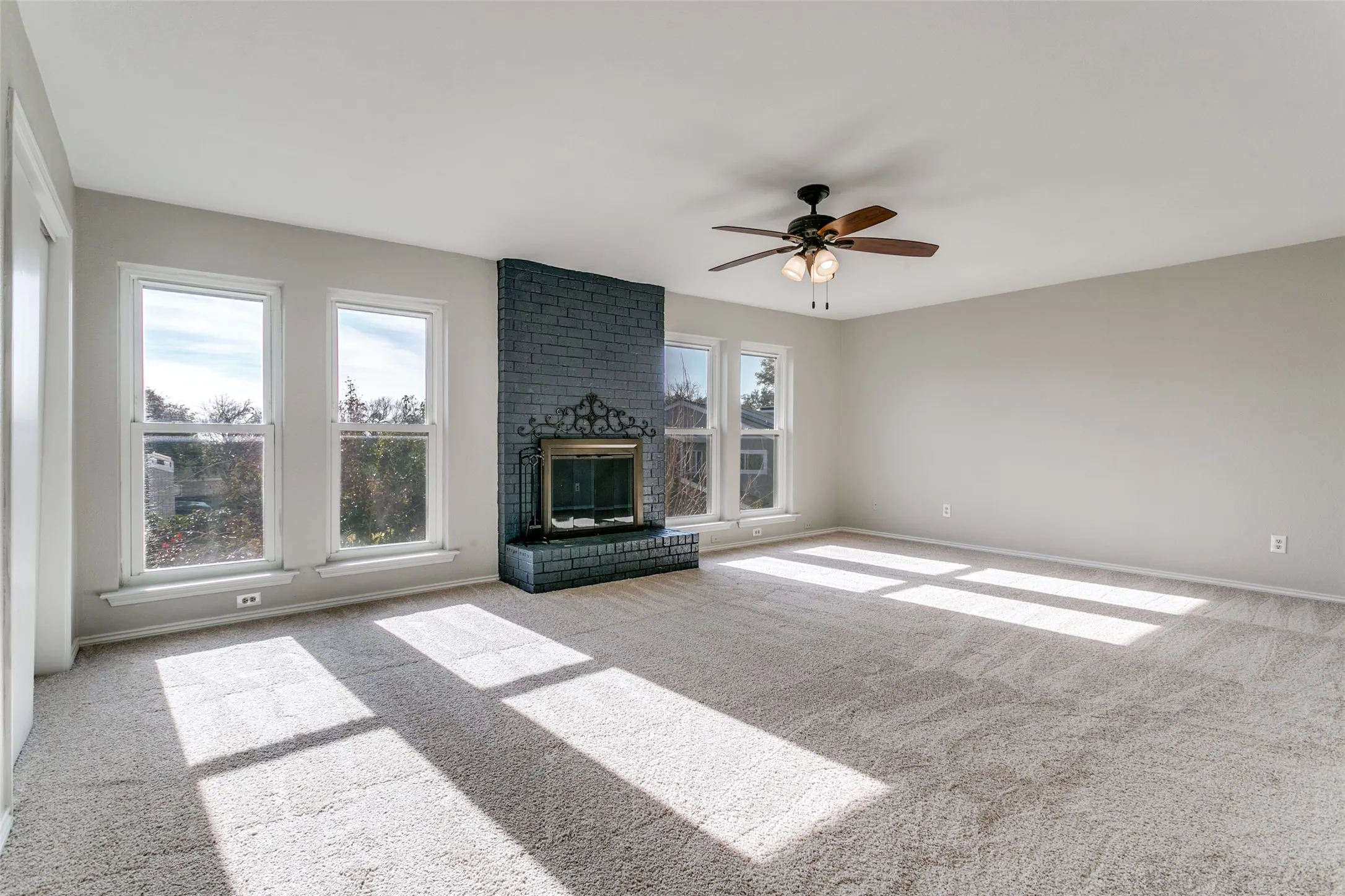 Spacious room featuring a dark brick fireplace, multiple windows, light neutral wall paint, and a ceiling fan with integrated lighting