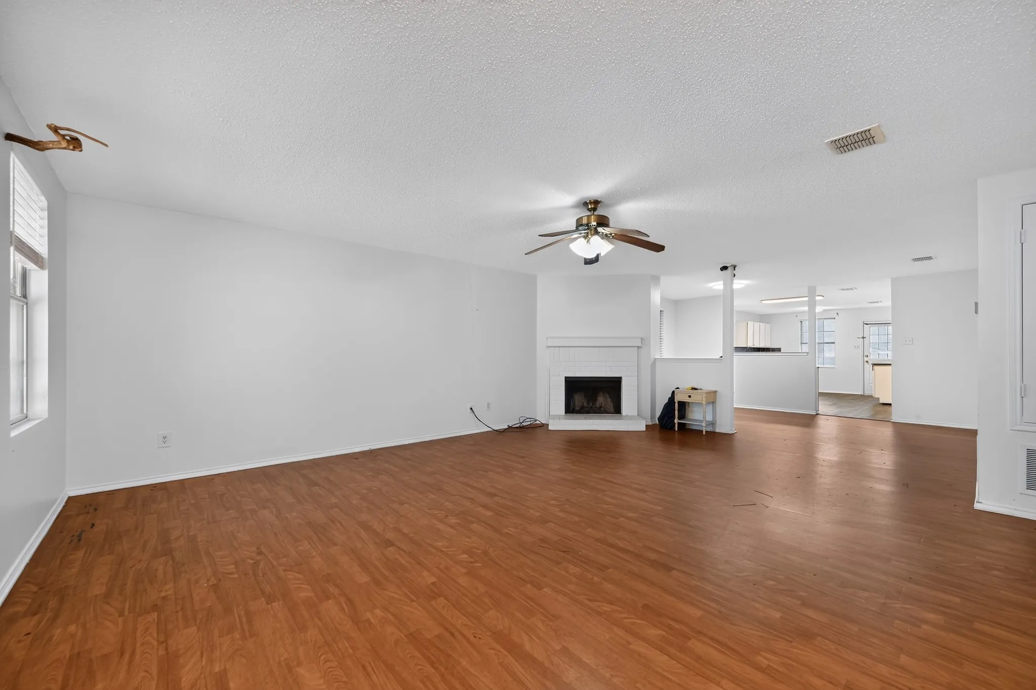 Expansive living area featuring wood-finish flooring, a white-painted brick fireplace with a mantel, and a ceiling fan