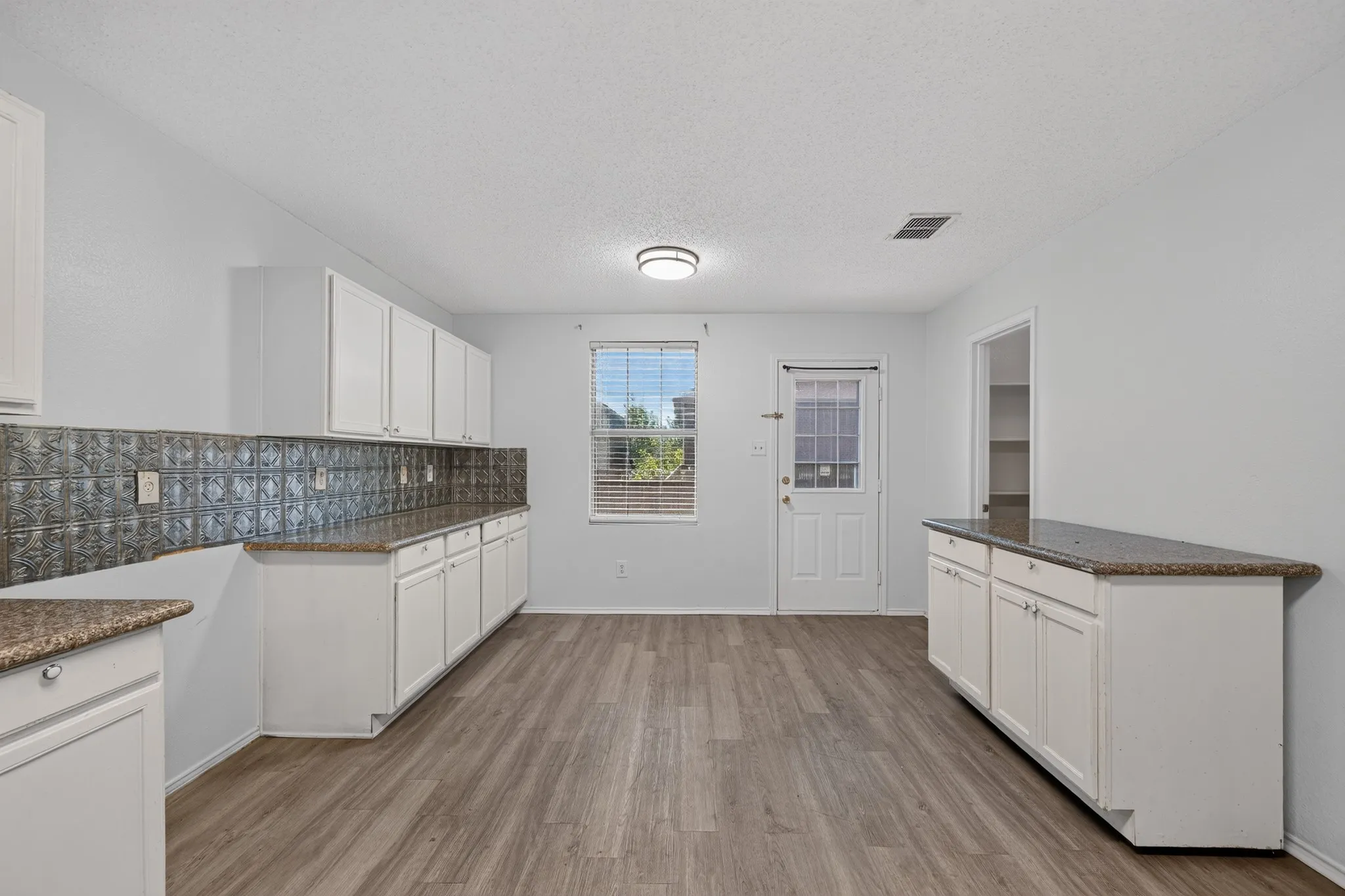 Spacious kitchen featuring extensive white cabinetry, speckled countertops, wood-finish flooring, a decorative tile backsplash, and ample natural light
