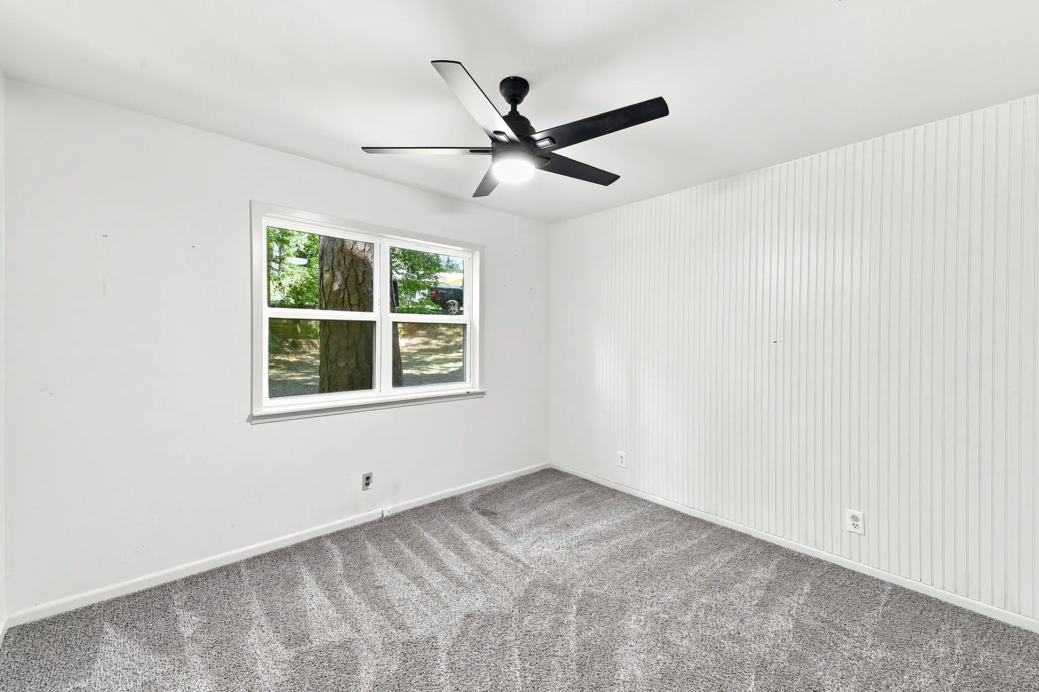 Bedroom featuring a single window overlooking exterior trees, a black ceiling fan with integrated lighting, a white beadboard accent wall, and gray patterned carpeting