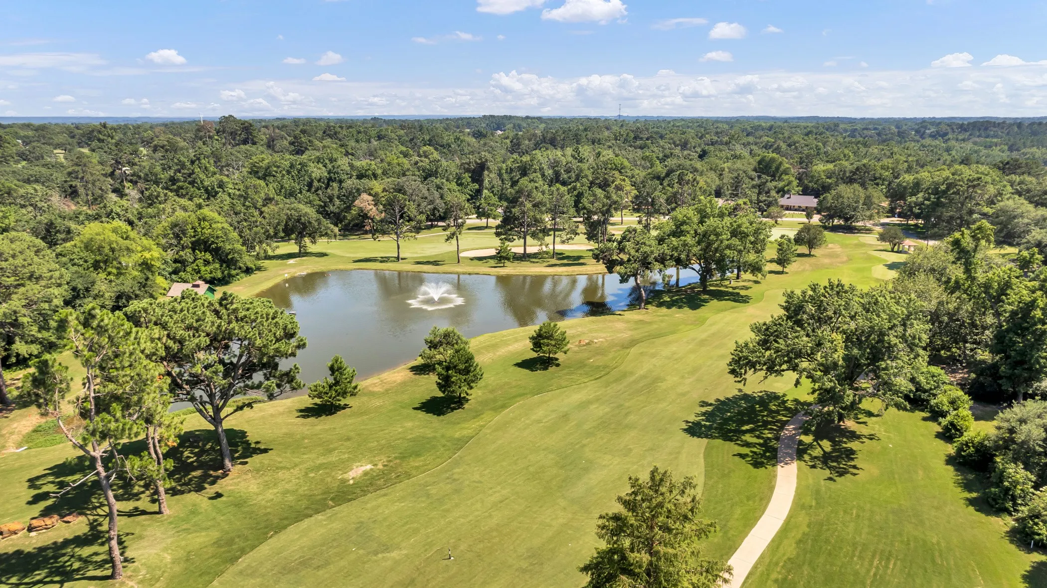 Expansive golf course property featuring a central pond with a fountain, mature trees, manicured greens, and a paved cart path