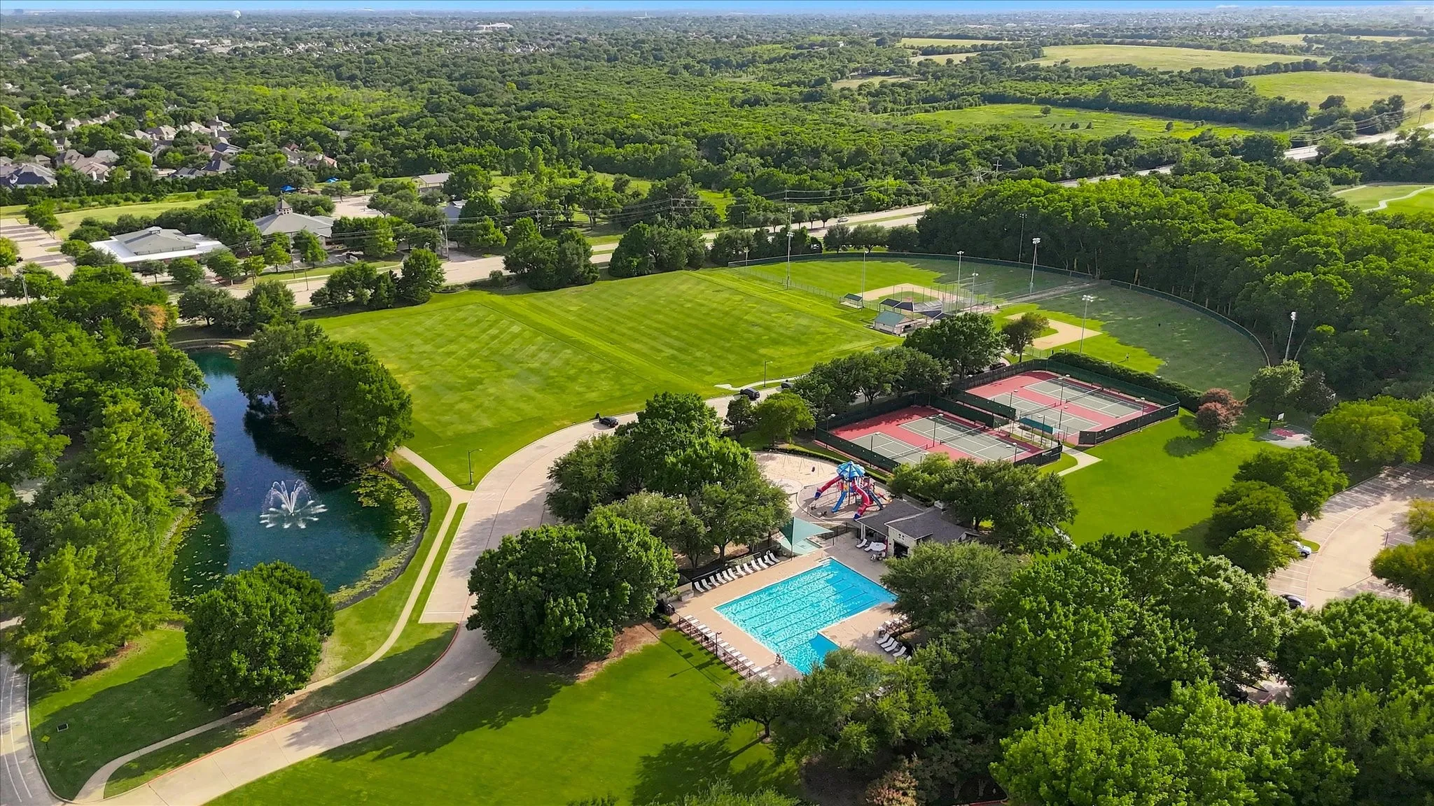 Expansive green spaces featuring a swimming pool, tennis courts, baseball fields, and a pond with a fountain