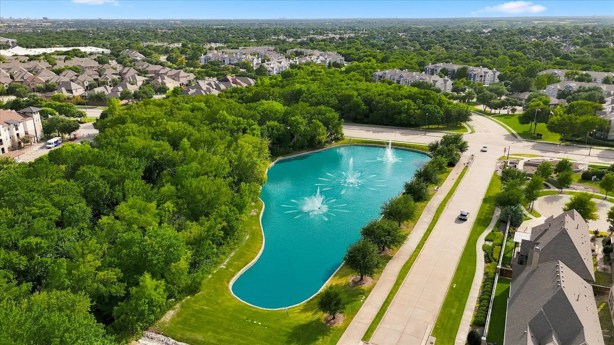 Expansive urban setting with a prominent pond featuring multiple fountains, surrounded by mature trees and residential structures