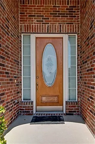 Brick exterior facade with a wood-finish entry door featuring an oval frosted glass insert, flanked by two sidelights with horizontal blinds, all set on a concrete porch